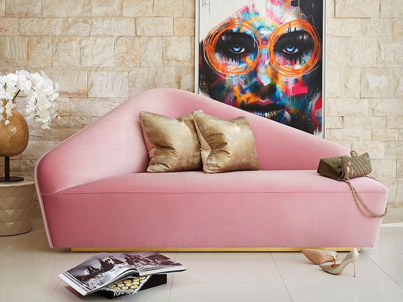 Modern living room with a pink velvet sofa, gold pillows, colorful abstract face artwork, white flowers in a vase, and a fashion magazine on the floor.