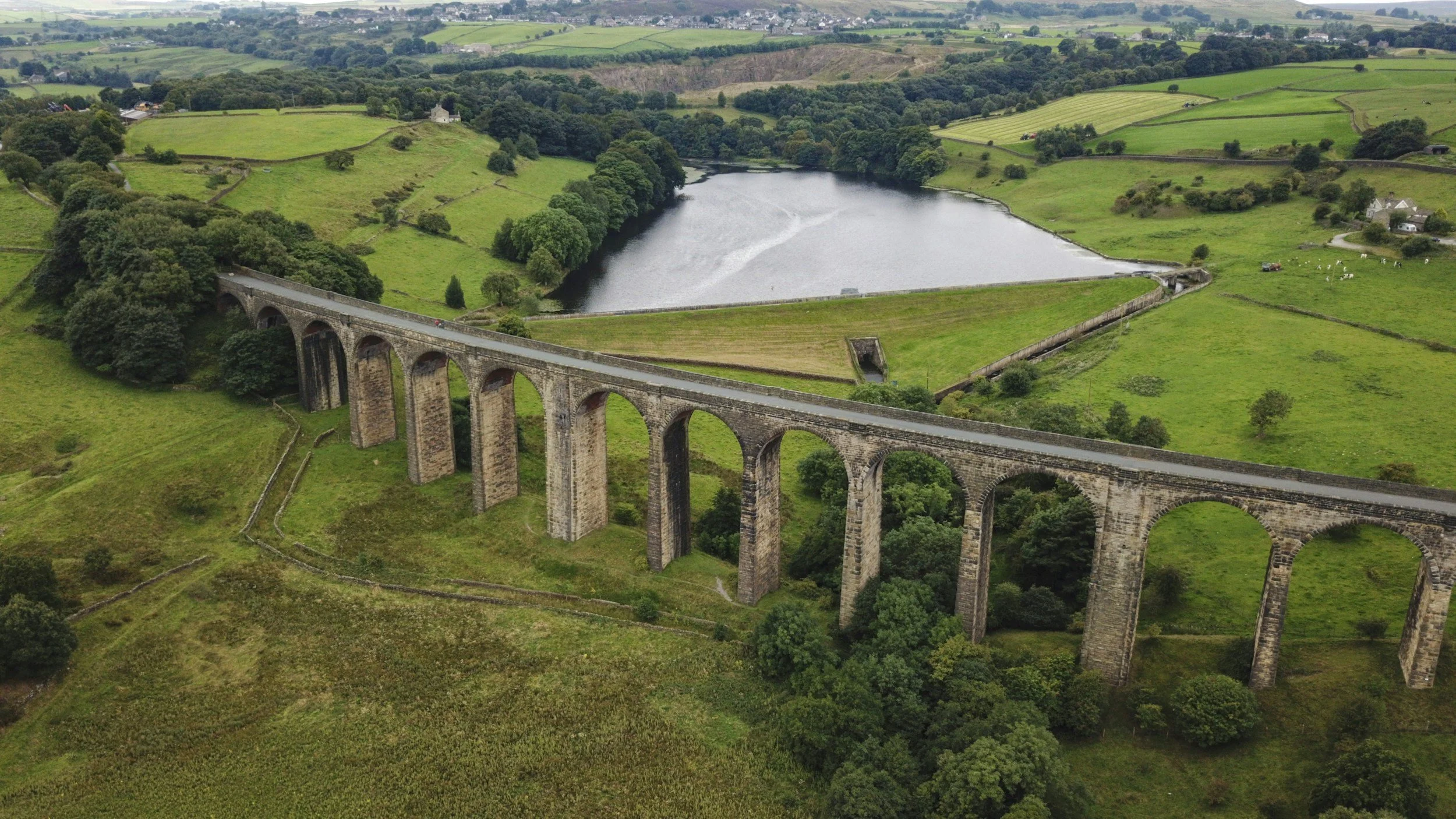 Aerial view of a stone railway viaduct with multiple arches crossing green farmland and a reservoir in a rural landscape.