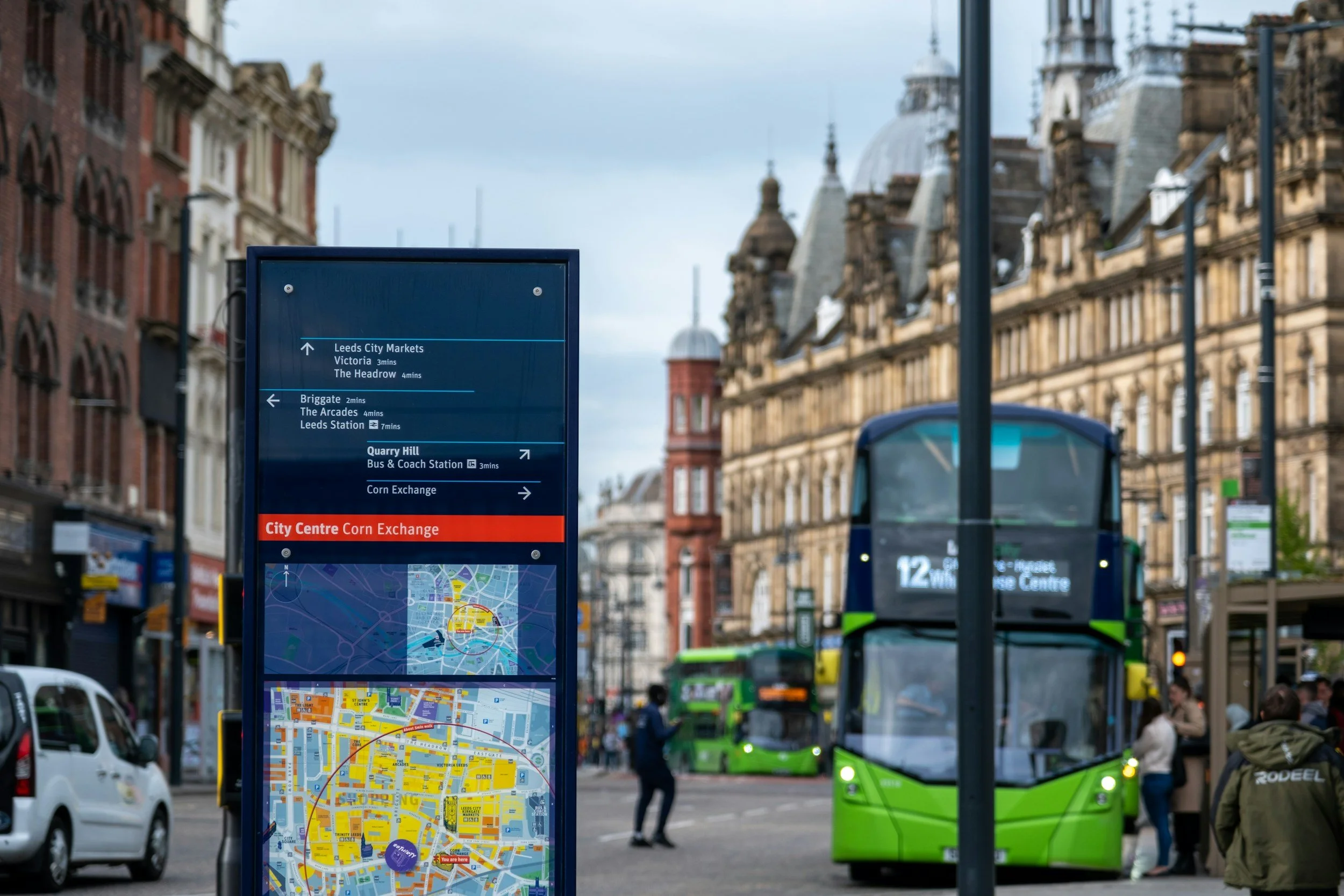 City street scene with a bus stop info board, green double-decker bus, and historic buildings in the background.