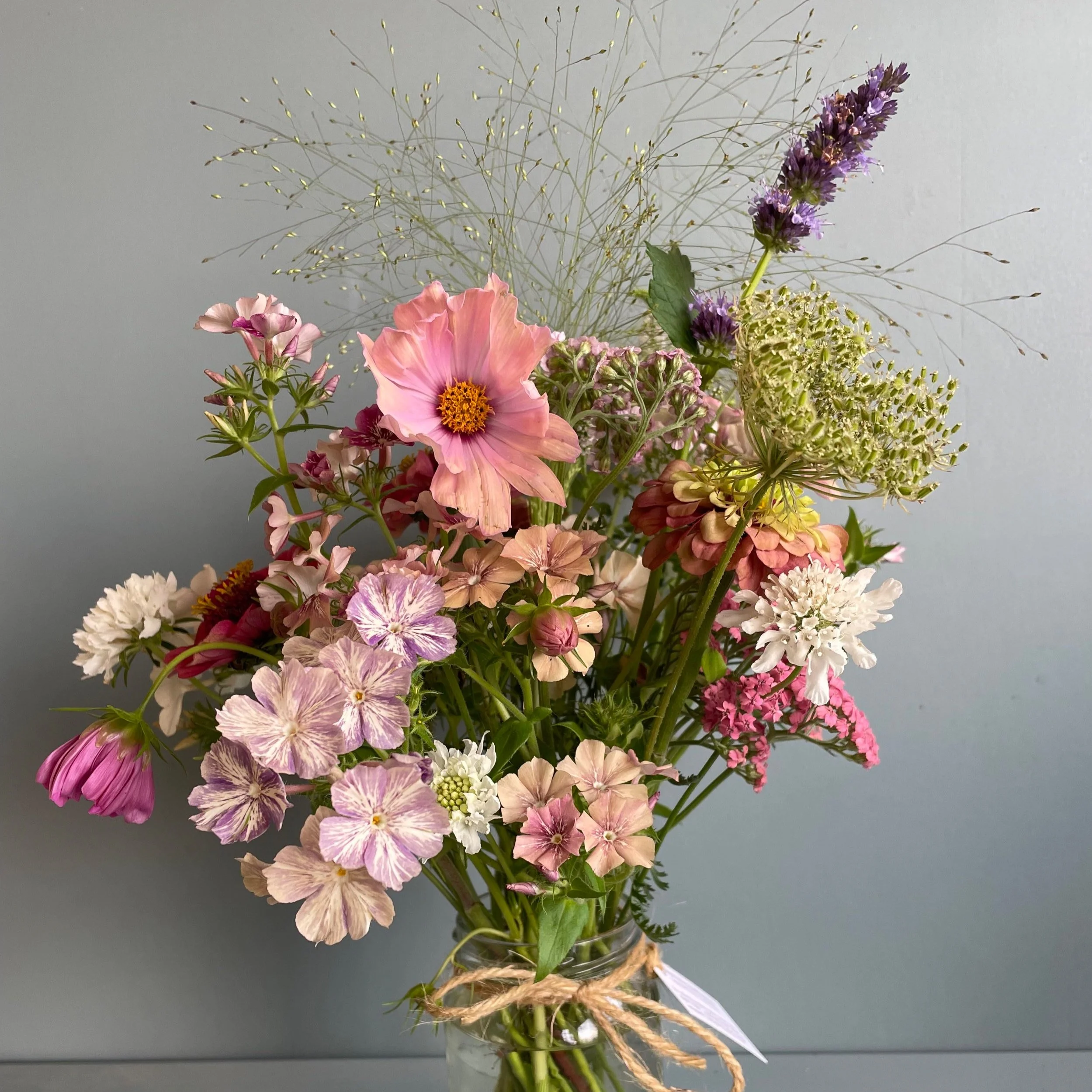 A glass vase filled with a colorful assortment of fresh flowers, including pink, purple, white, and peach blooms, with some greenery and delicate filler flowers, against a plain gray background.