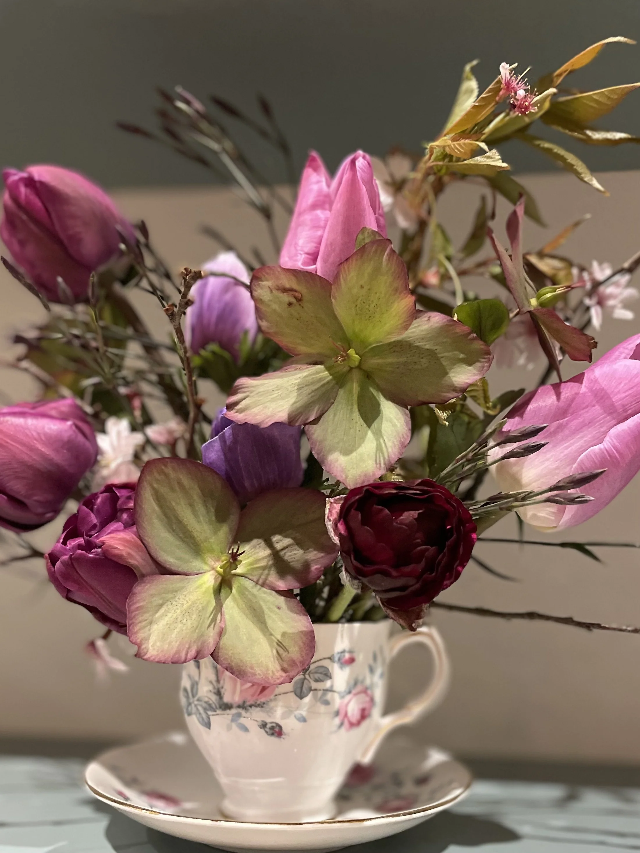 A floral teacup with a matching saucer holding a bouquet of mixed flowers, including tulips and hellebores