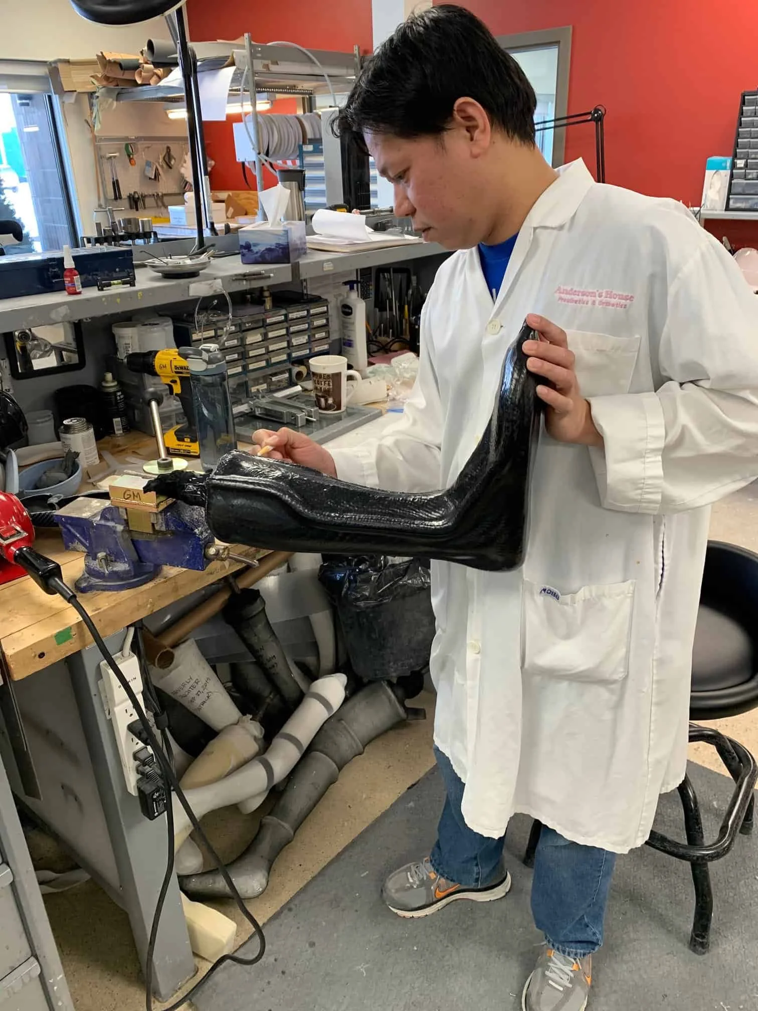 A person in a white lab coat working in a workshop, holding a black prosthetic arm and using a small tool at a workbench surrounded by various tools and equipment.