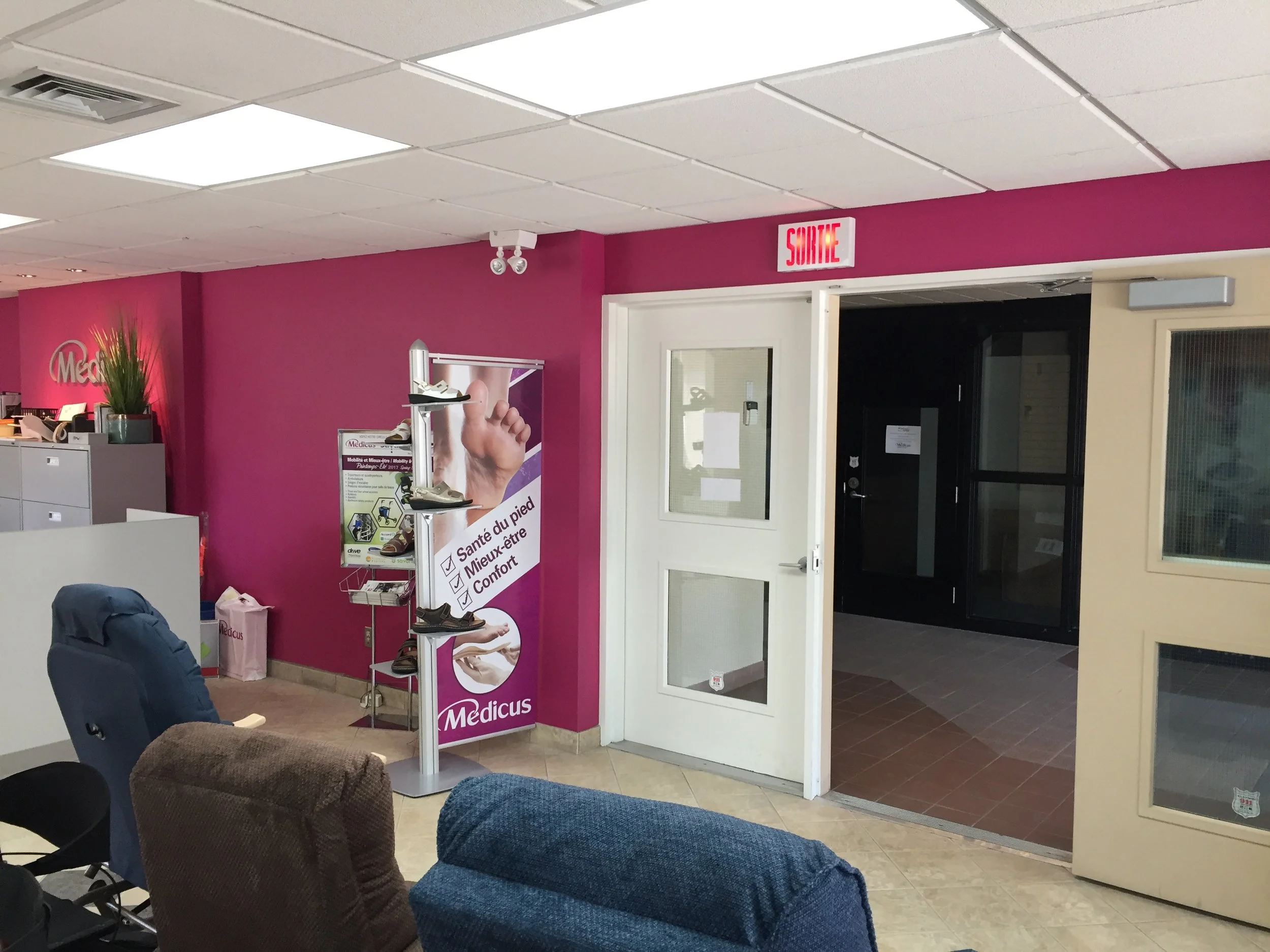 Intérieur d'une clinique médicale avec des murs roses, des chaises et un présentoir à chaussures près d'une porte blanche avec un panneau « sortie ».
