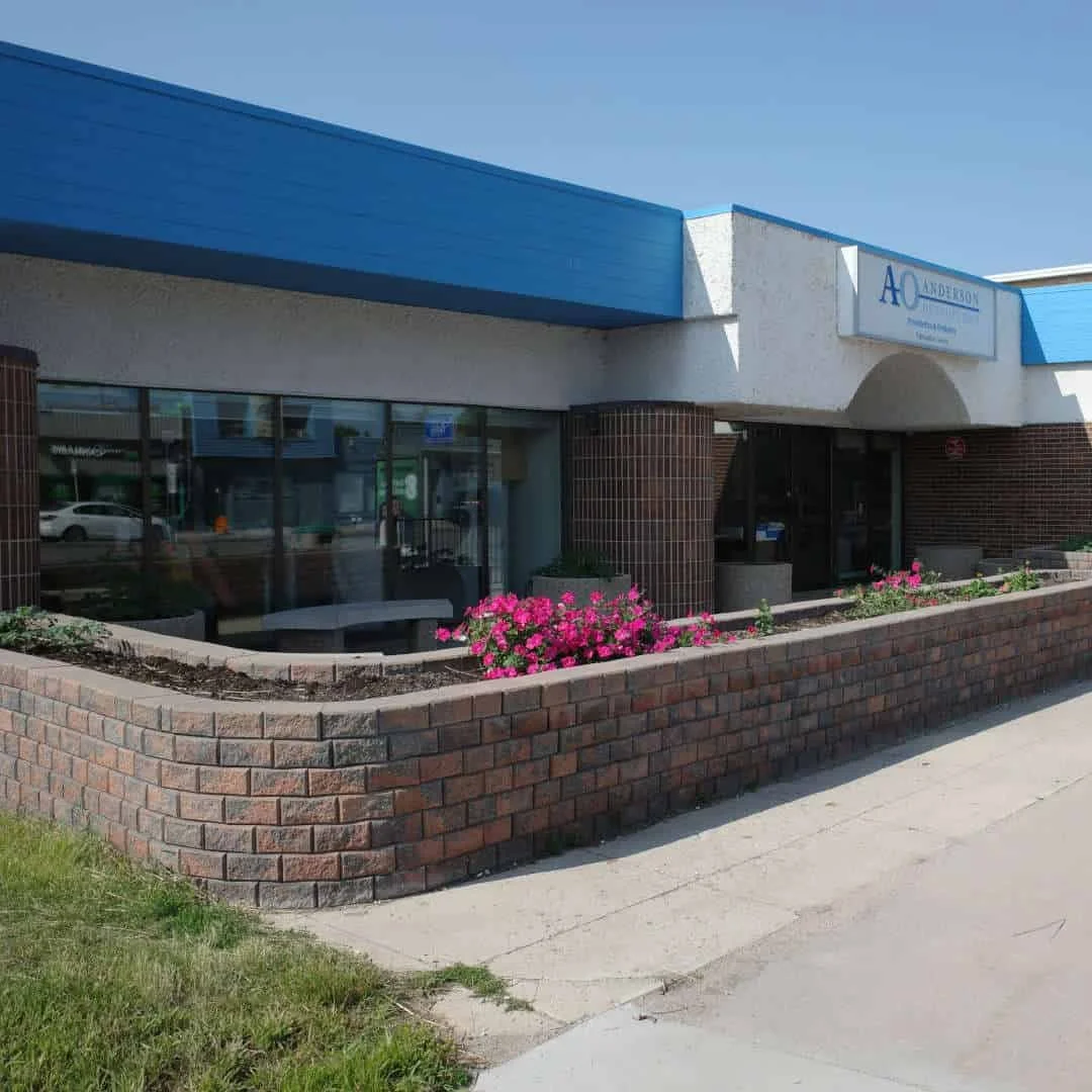 Extérieur d'un bâtiment avec de grandes fenêtres, un mur de briques et une enseigne blanche et bleue sur laquelle on peut lire « AO Anderson ». Des fleurs roses sont plantées dans un parterre surélevé en briques devant le bâtiment.