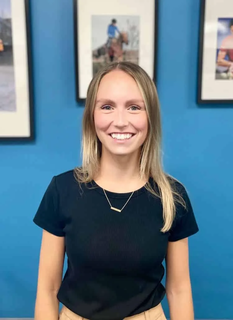 Une jeune femme blonde, souriante, vêtue d'une chemise noire et d'un collier délicat, debout devant un mur bleu orné de photographies encadrées.
