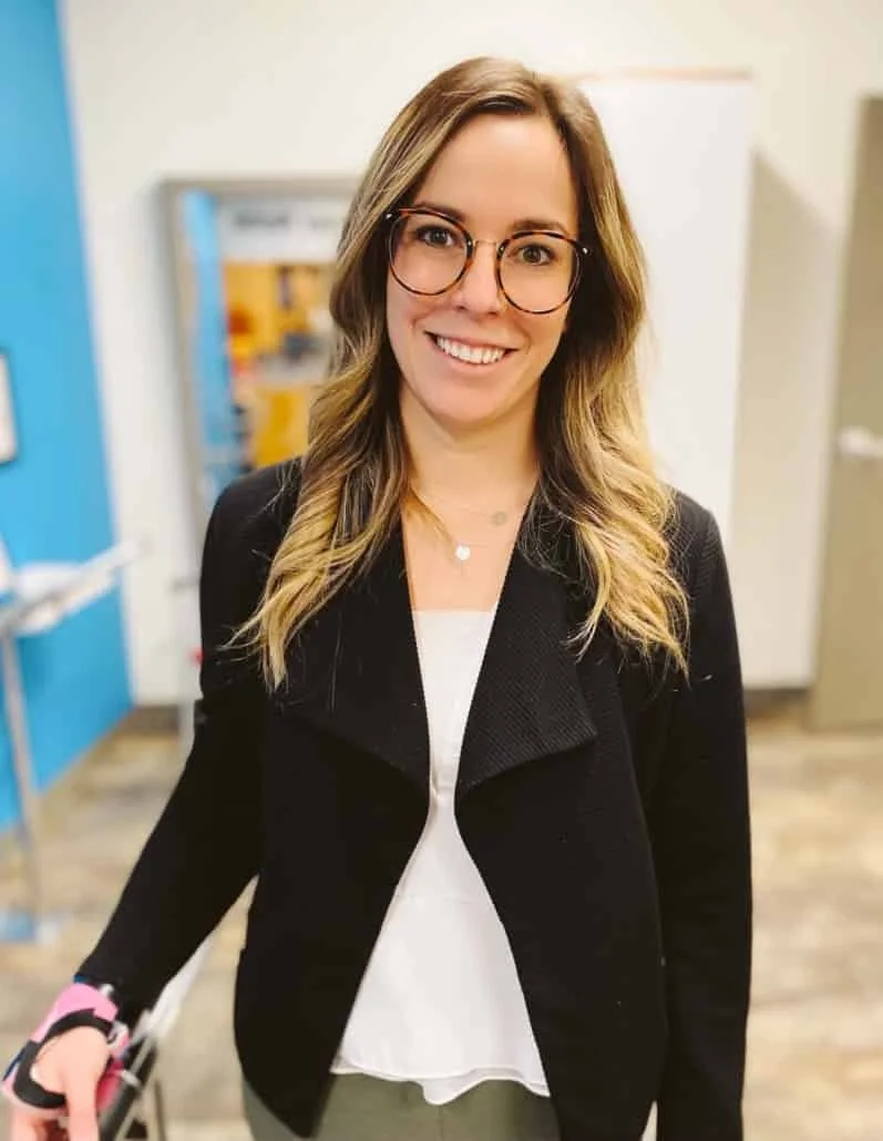 Une jeune femme aux longs cheveux blonds ondulés, portant des lunettes rondes, un blazer noir et un chemisier blanc, souriant alors qu'elle se tient debout à l'intérieur.