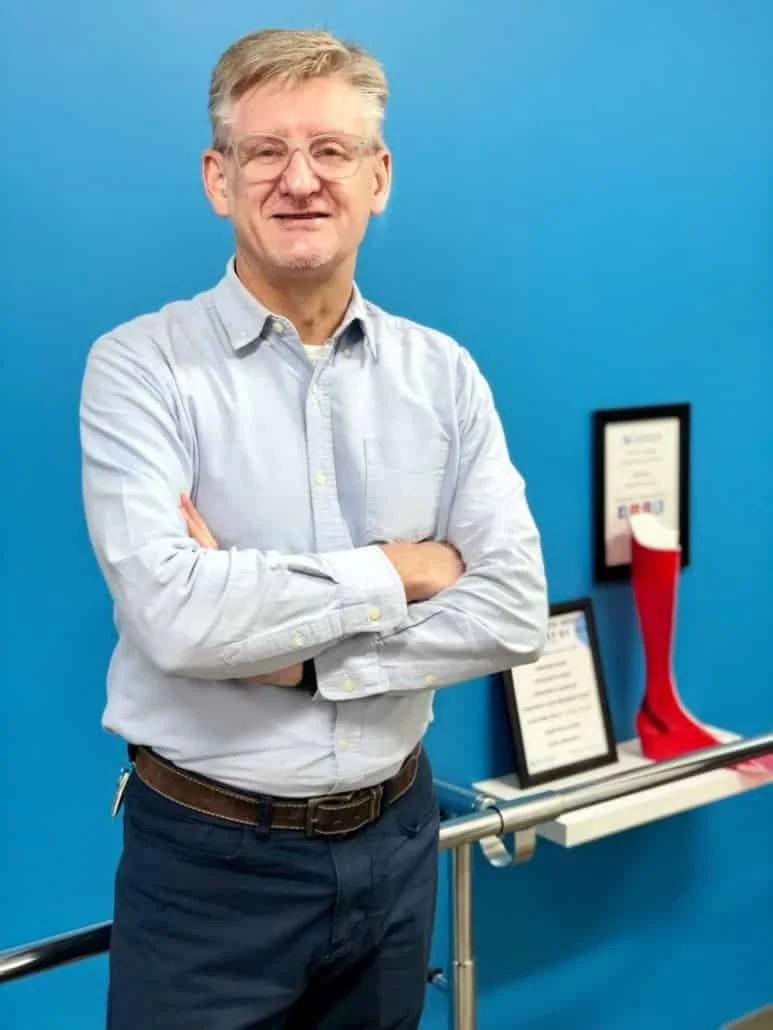 A man with glasses, gray hair, and a light blue button-up shirt standing with arms crossed in front of a blue wall. A framed certificate and a red Christmas stocking are on a table behind him.
