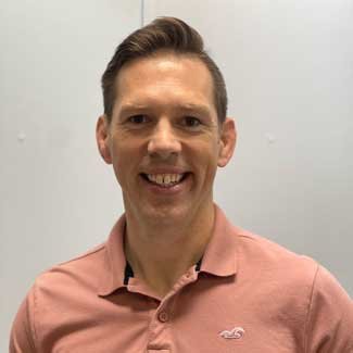 A young man smiling in front of a plain light-colored wall, wearing a pink collared shirt.