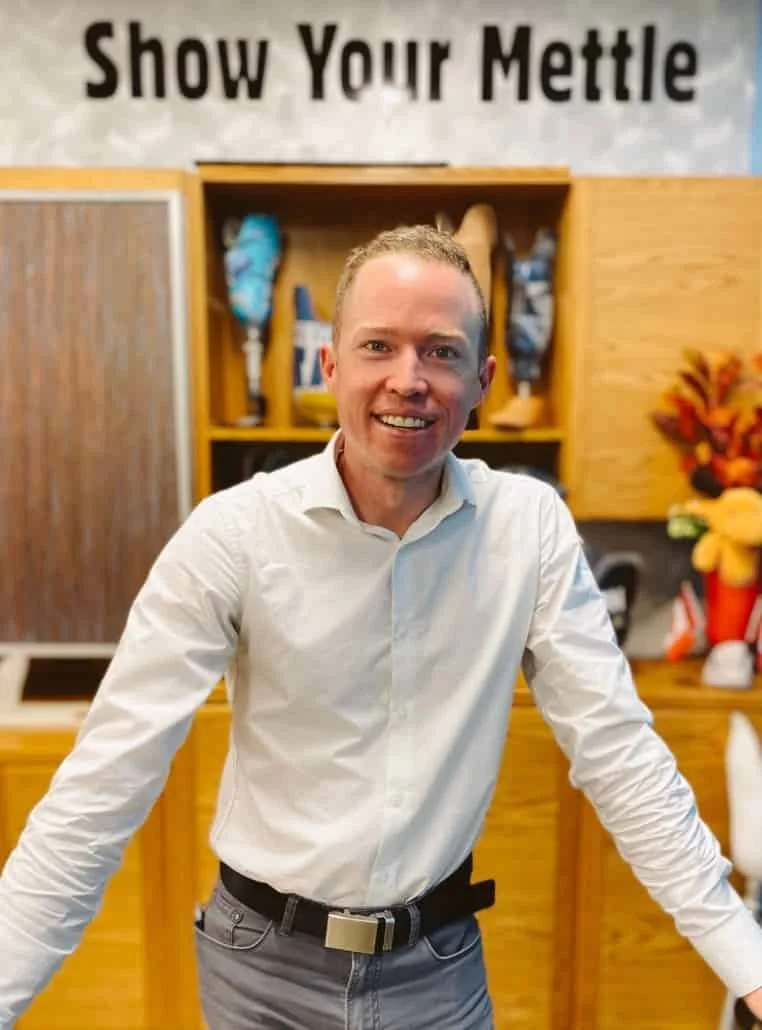 Un homme souriant vêtu d'une chemise blanche et d'un pantalon gris, debout devant une armoire en bois contenant des objets décoratifs et une pancarte sur laquelle on peut lire « Montrez ce que vous valez ».