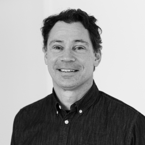 Portrait en noir et blanc d'un homme souriant aux cheveux courts et ondulés, vêtu d'une chemise à col.