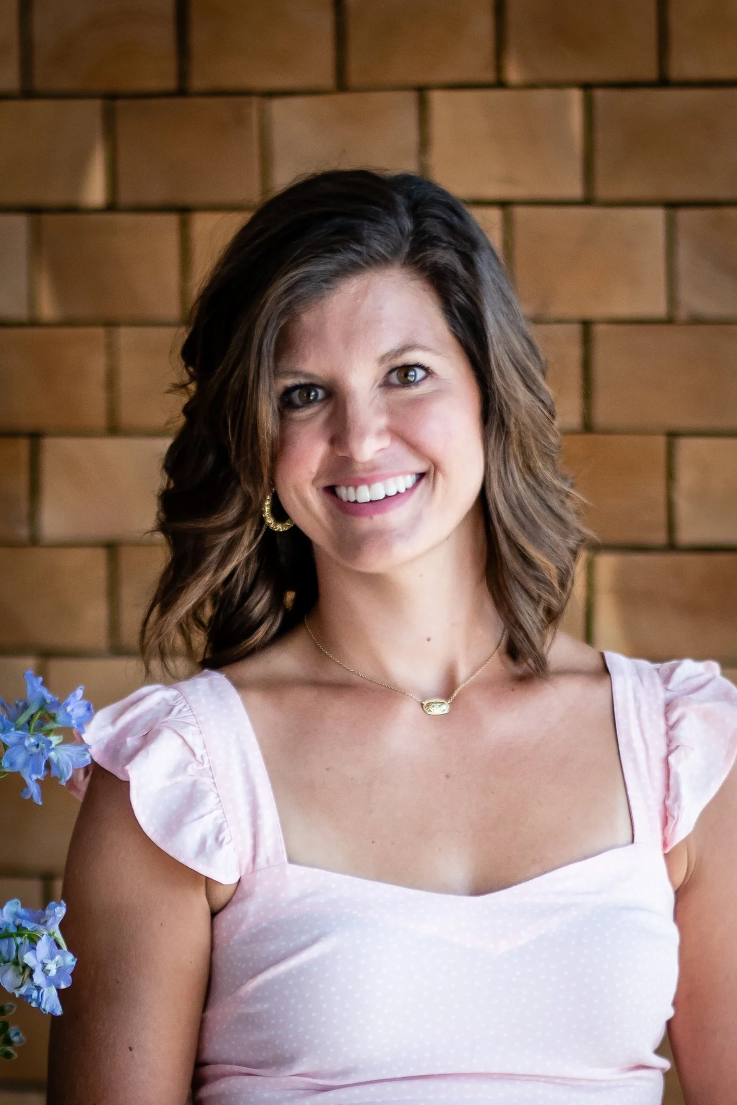 A woman with shoulder-length brown hair smiling in front of a brick wall, wearing a light pink dress with puffy sleeves, gold earrings, and a gold necklace.