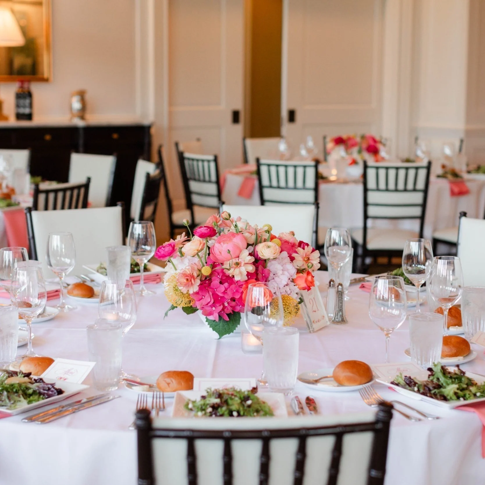 A beautifully decorated banquet table with a vibrant pink and white floral centerpiece, set for a special event with plates, silverware, glasses, and bread rolls.