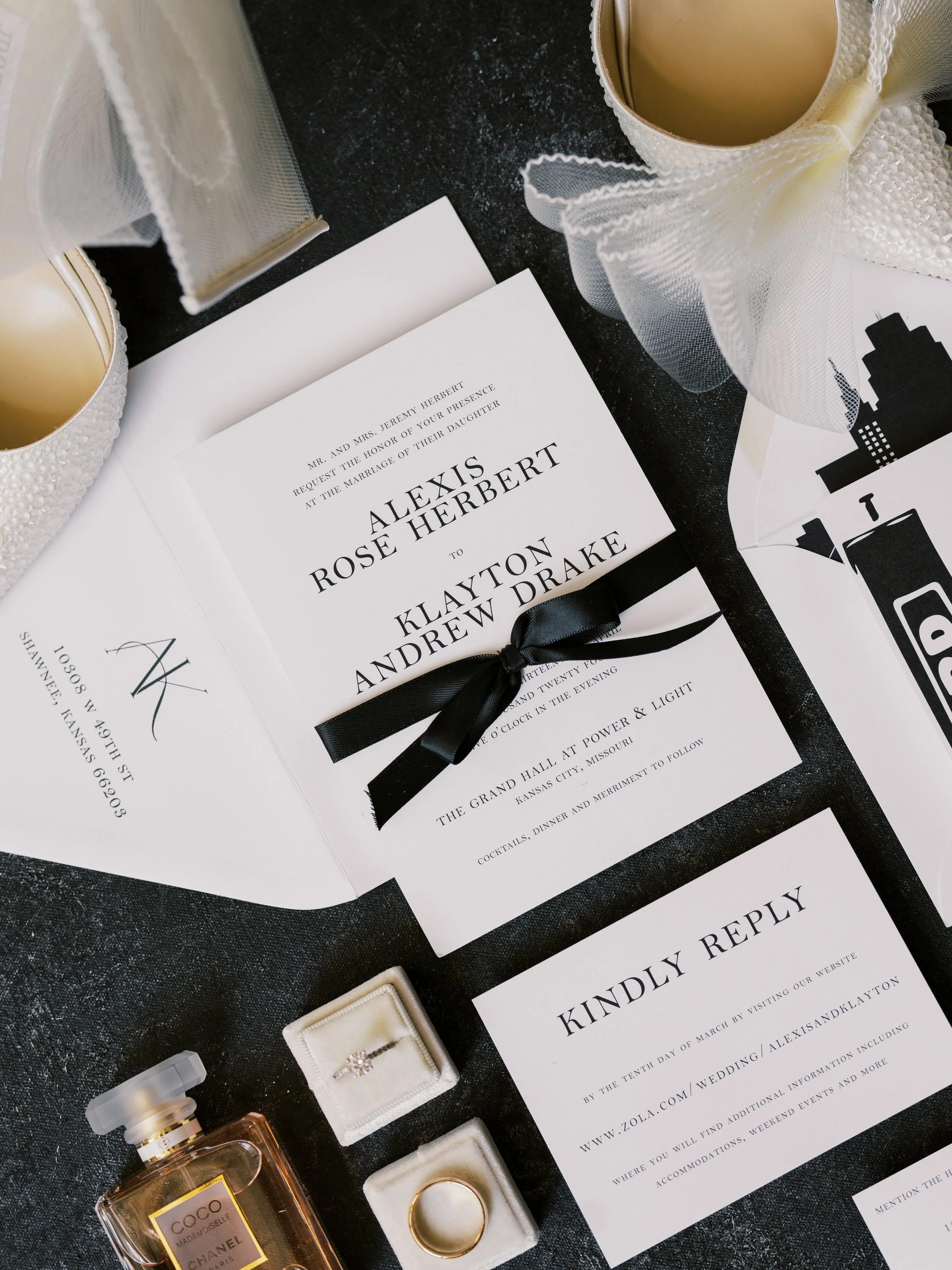 Wedding invitation with black ribbon, wedding ring, engagement ring, perfume, candles, and reply card on dark fabric background.