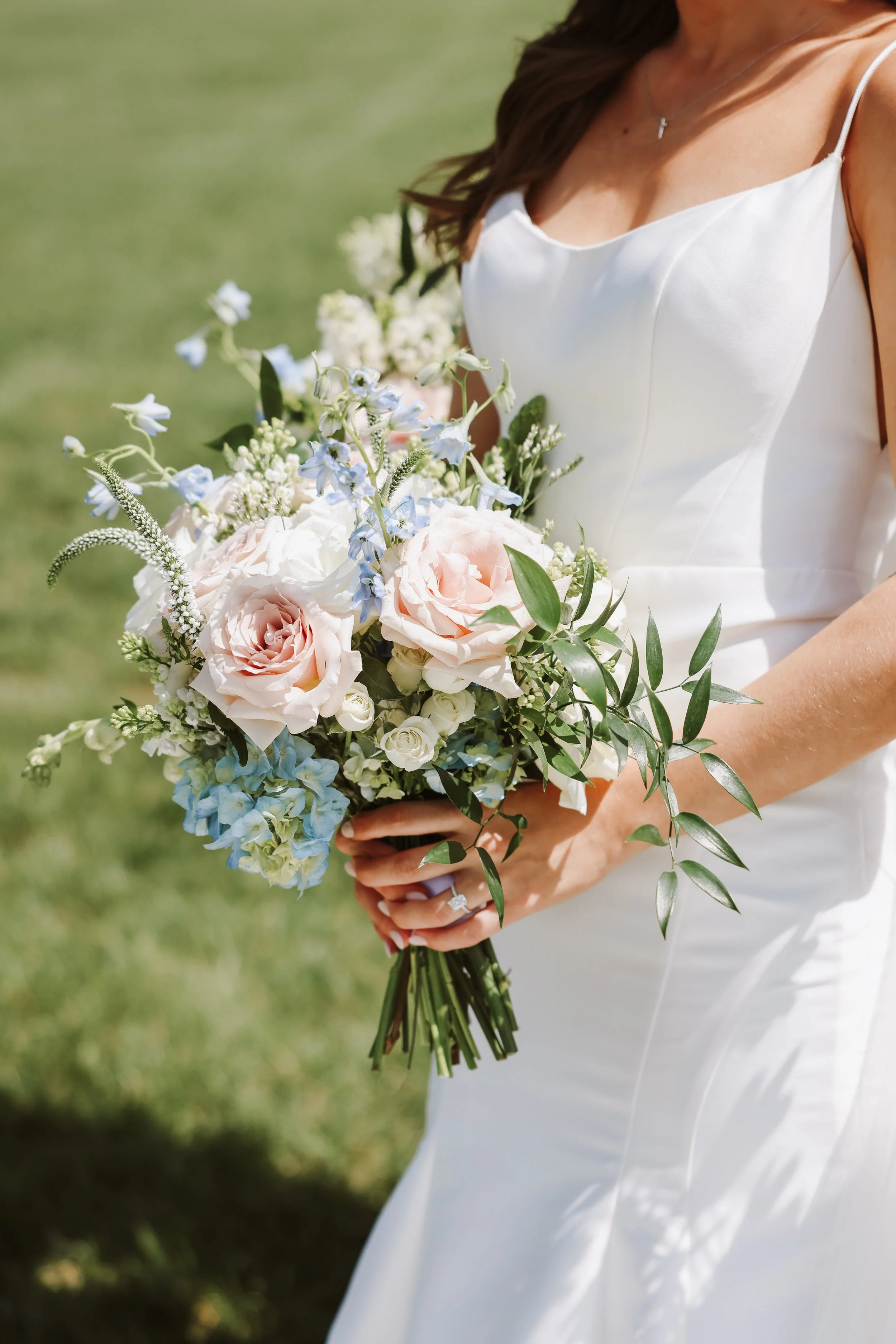 Bride holding a bouquet of light pink roses, white, and blue flowers with green leaves outdoors