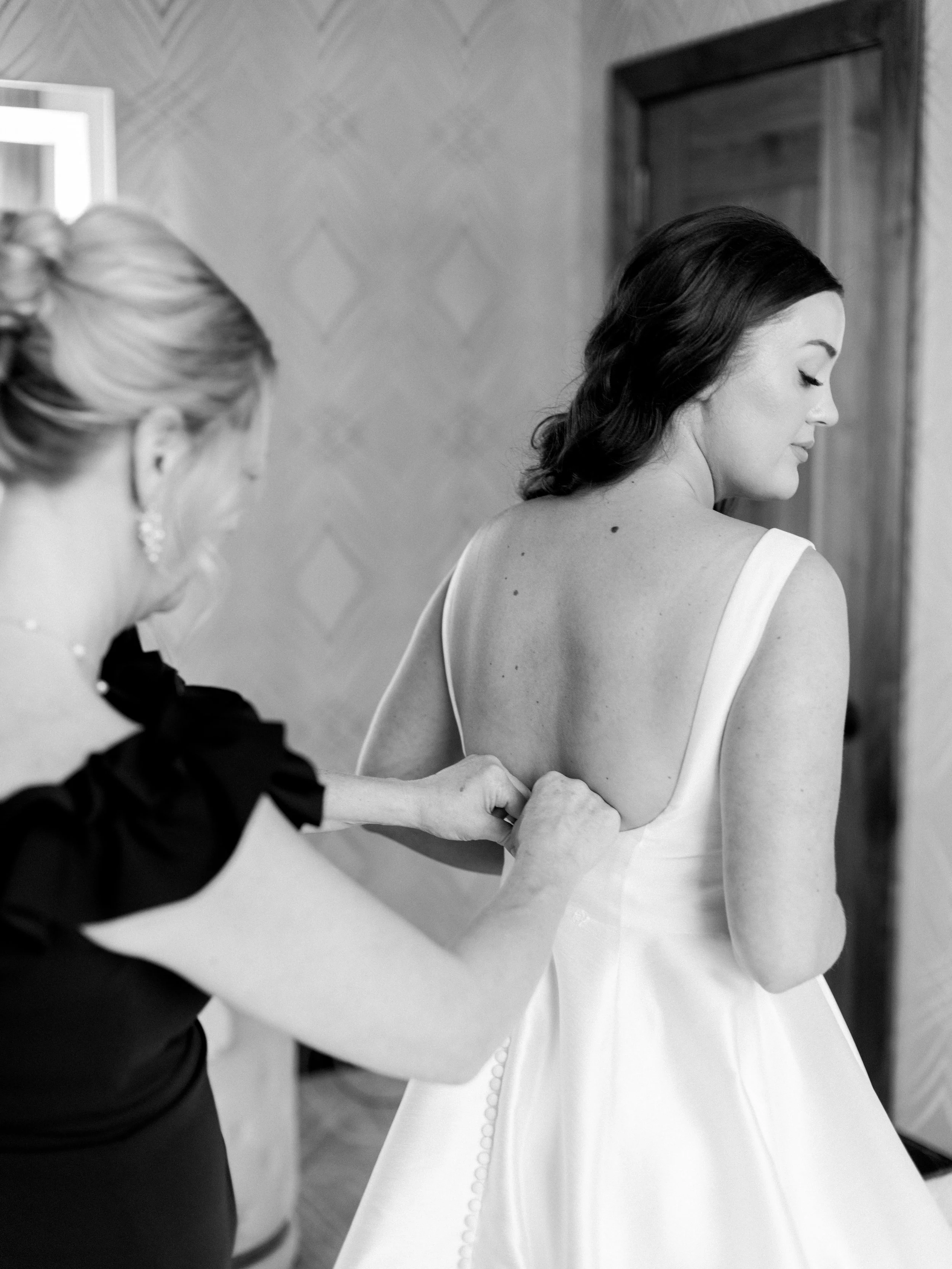 A woman in a wedding dress has the back of her dress buttoned by another woman in an elegant black dress, in a room with patterned walls and wooden door.