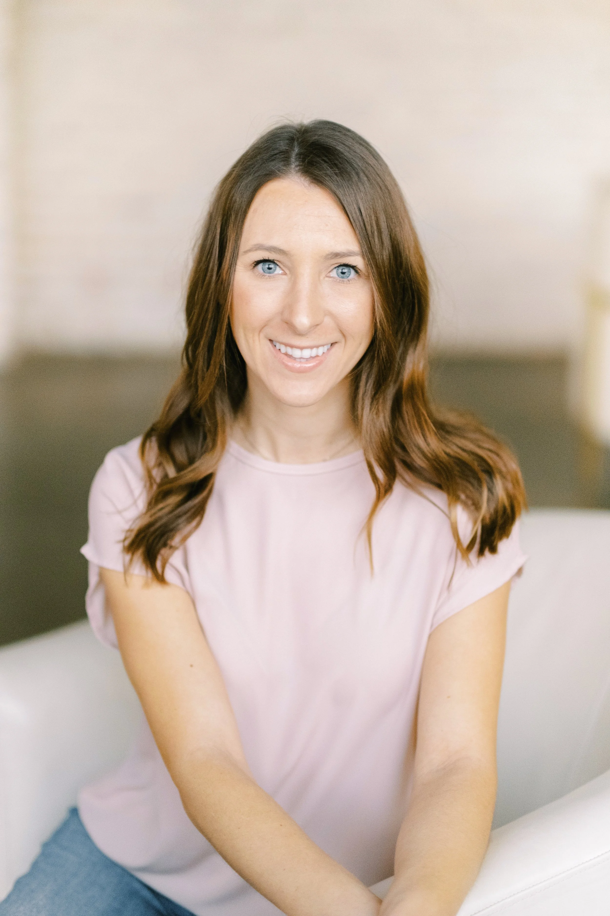A smiling woman with wavy brown hair and blue eyes sitting on a white couch in a bright room.