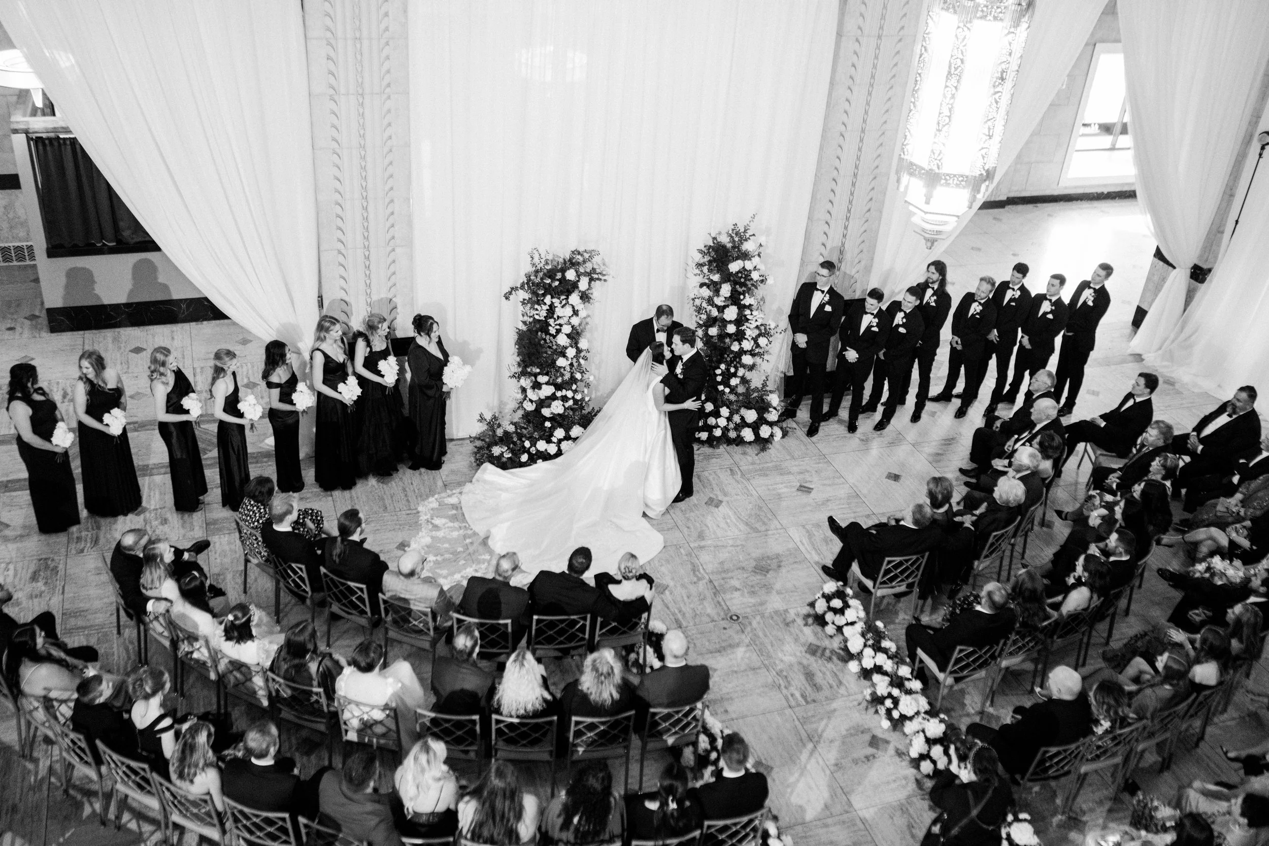 Black and white photo of a wedding ceremony with the bride and groom kissing in front of an altar, surrounded by family and friends seated in rows, with bridesmaids and groomsmen standing on either side.