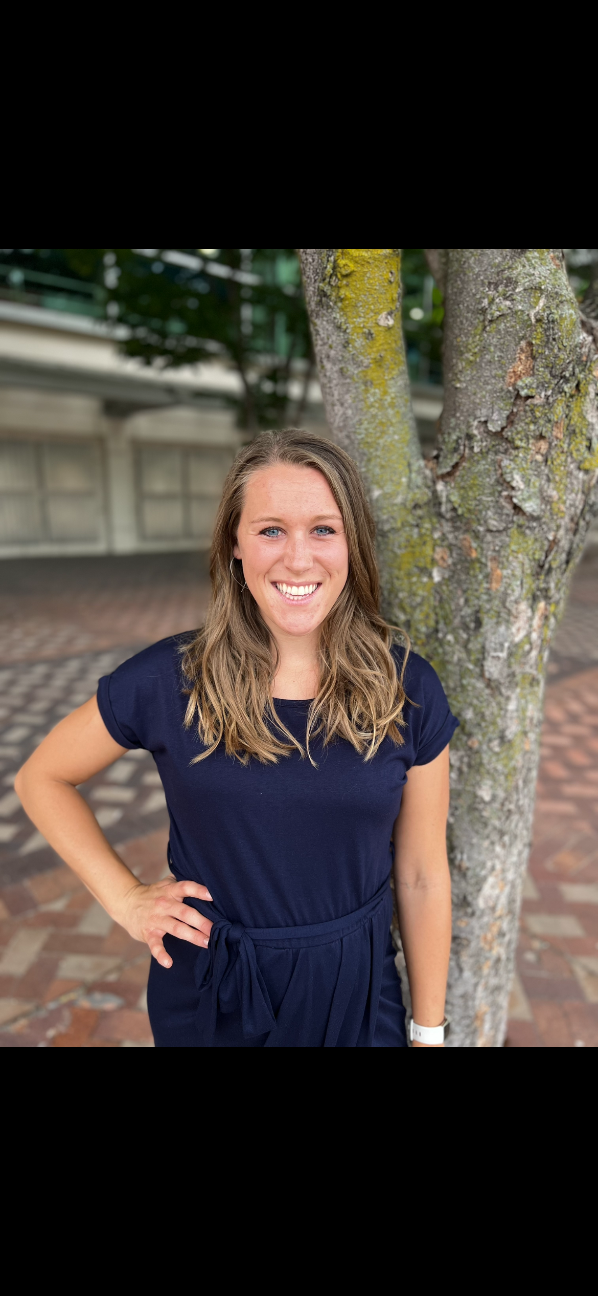 A smiling woman with shoulder-length wavy light brown hair, wearing a navy blue dress with a waist tie, standing outdoors next to a tree with moss on the bark.