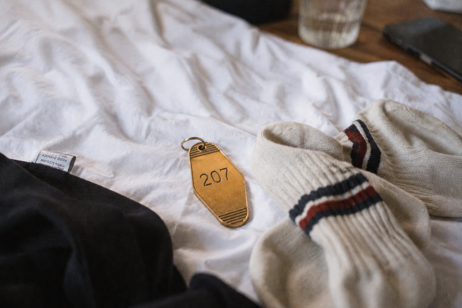 Ein Klebezettel mit der Nummer 207 liegt auf einem weißen Bettlaken. Daneben sind eine schwarze Kleidung, eine weißen Socke mit roten und schwarzen Streifen, eine Tasse und ein Smartphone sichtbar.