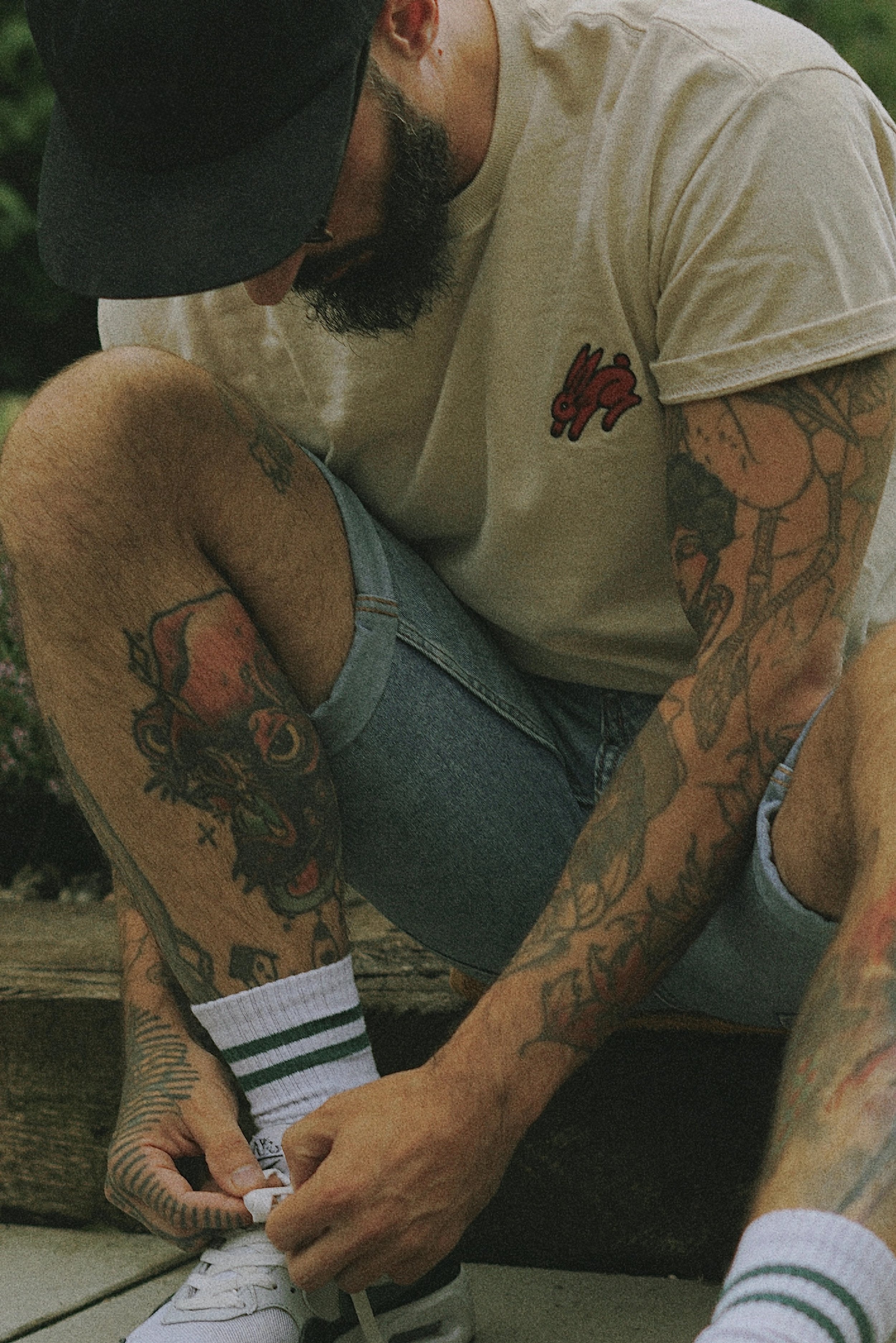 Ein Mann mit Tattoos, trägt ein beige T-Shirt und einen schwarzen Cap, bindet seine weißen Sneakers. Er hat einen Bart und trägt eine Brille.