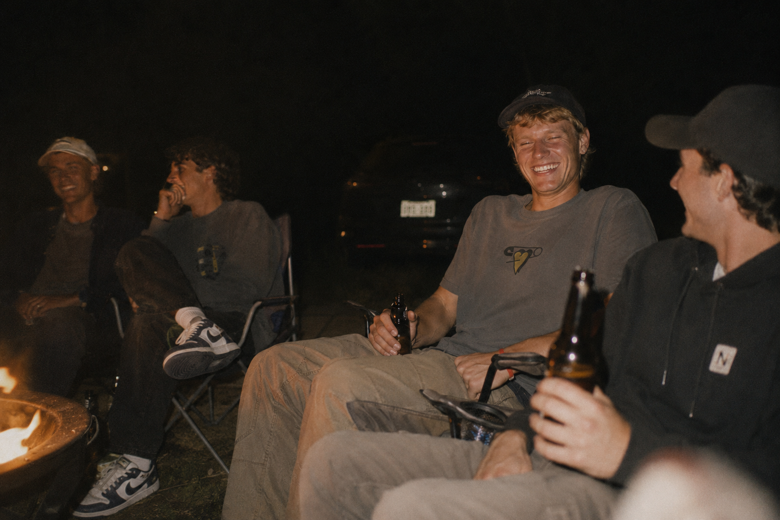Gruppe von Freunden, die abends um ein Lagerfeuer sitzen und lachen, Bier trinken und die Gesellschaft genießen.