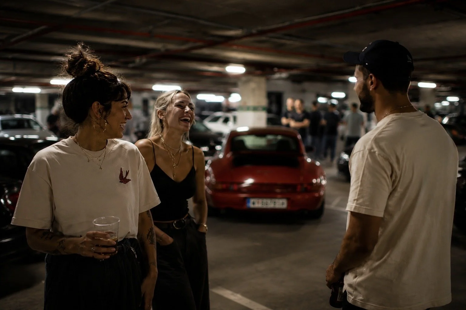 Drei Menschen lachen und unterhalten sich in einer Tiefgarage, zwei Frauen und ein Mann, im Hintergrund geparkte Autos.