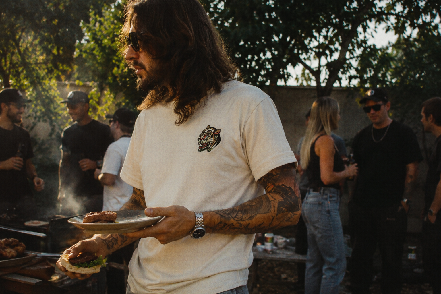 Ein Mann mit Tattoos und Sonnenbrille, der ein Sandwich hält, während er bei einem Grillfest im Freien steht. Im Hintergrund sind mehrere Menschen, die Drinks in der Hand halten, und es sind Bäume zu sehen.