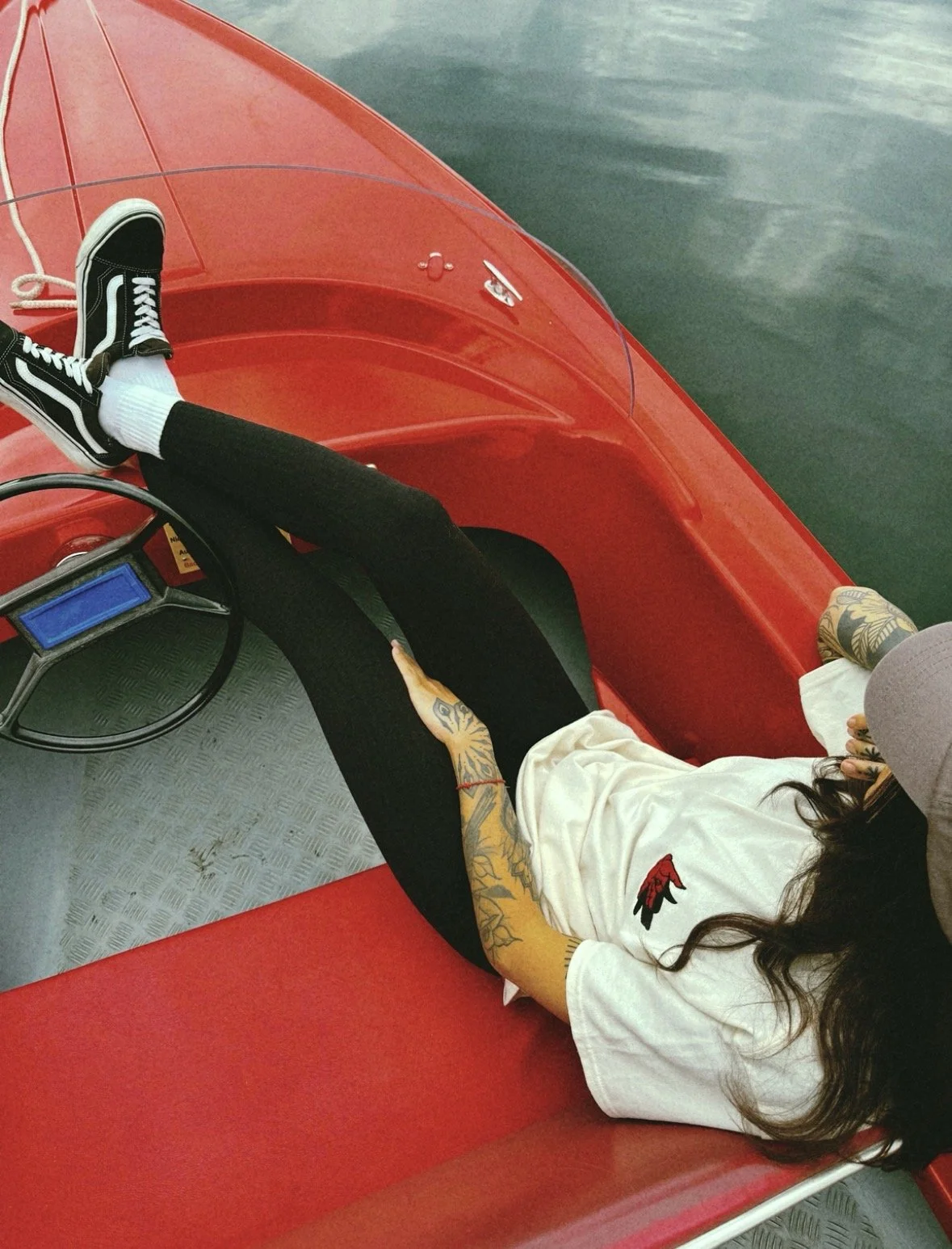 Person liegt entspannt in einem roten Elektroboot auf dem Wasser, trägt schwarze Leggings, weiße Socken und Vans-Schuhe, hat Tattoos am Arm und trägt ein weißes T-Shirt mit rotem und schwarzem Logo.