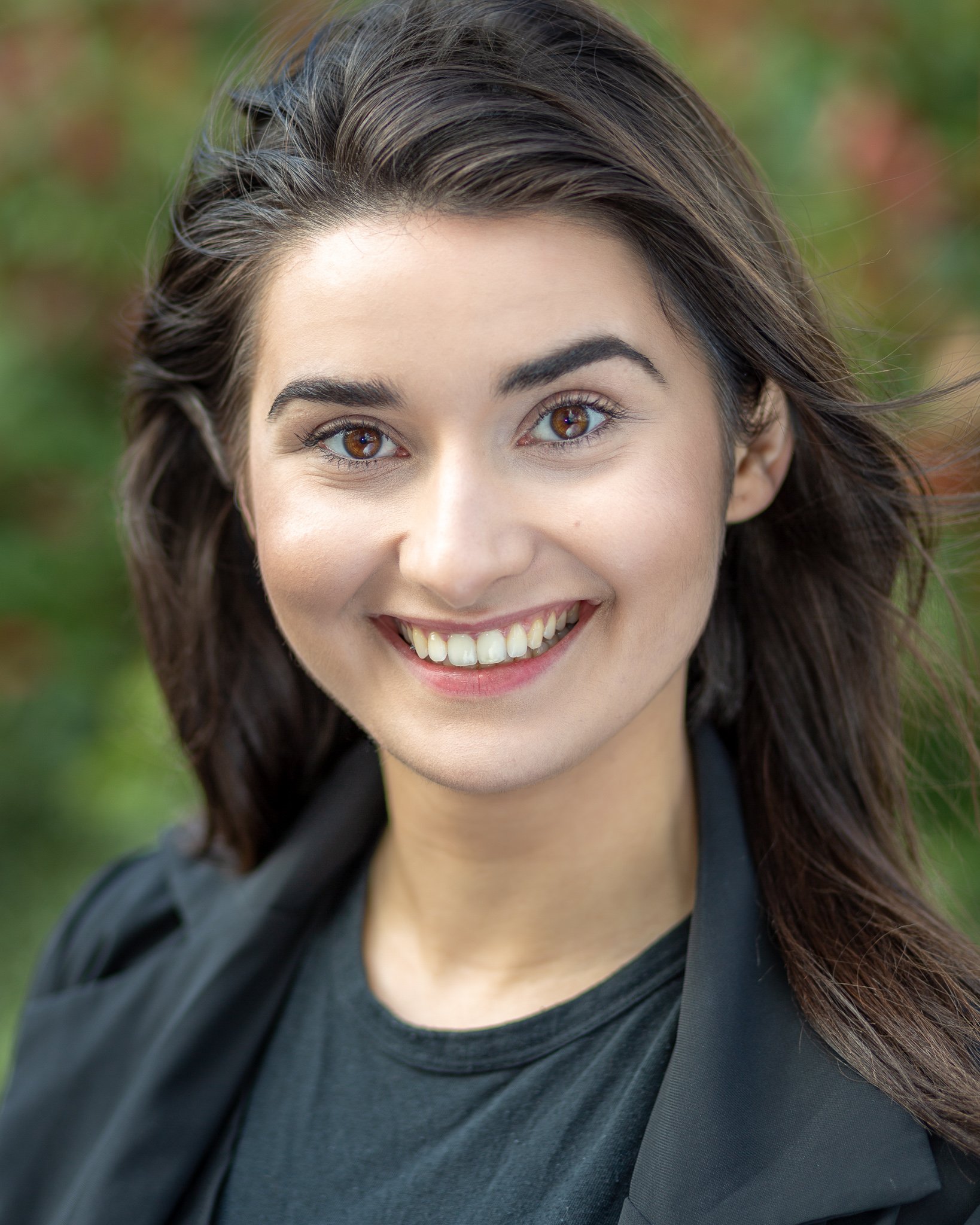 Smiling person with long dark hair, wearing a dark blazer and shirt, against a blurred green background.