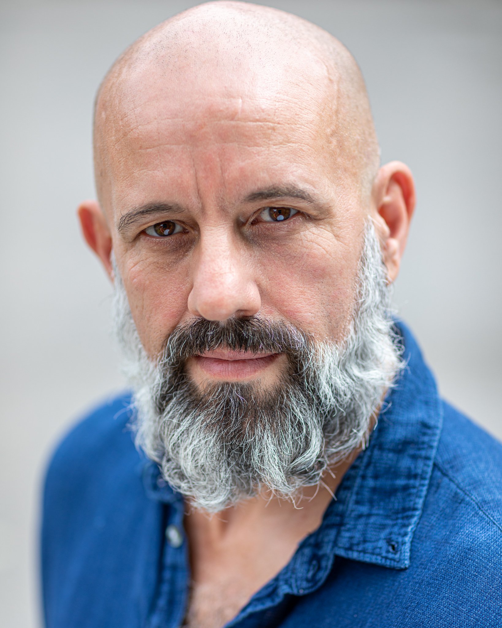 Bald man with a gray beard wearing a blue shirt, looking at the camera