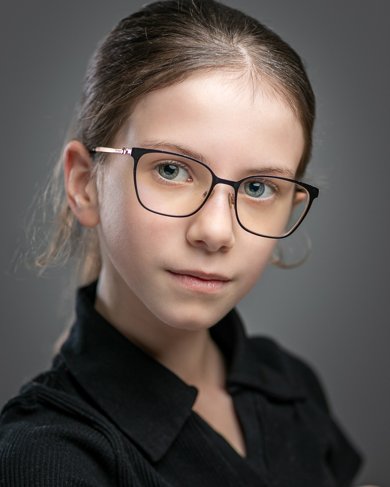 Young girl with glasses, wearing a dark shirt, looking forward with a soft expression.
