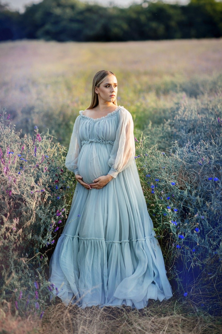 Schwangere Frau in einem blauen Kleid steht in einem Blumenfeld.