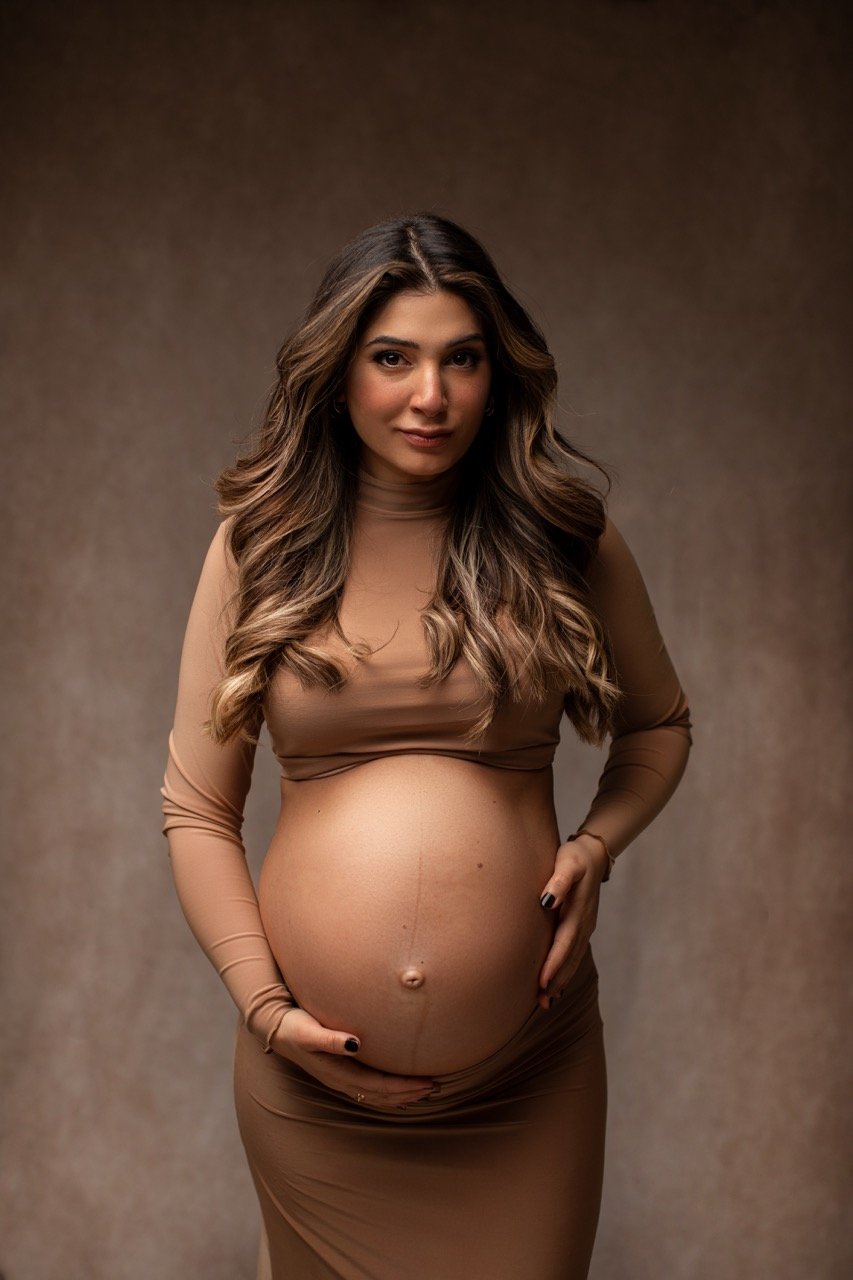 Eine schwangere Frau in einem beigen Kleid hält ihren Bauch mit beiden Händen.