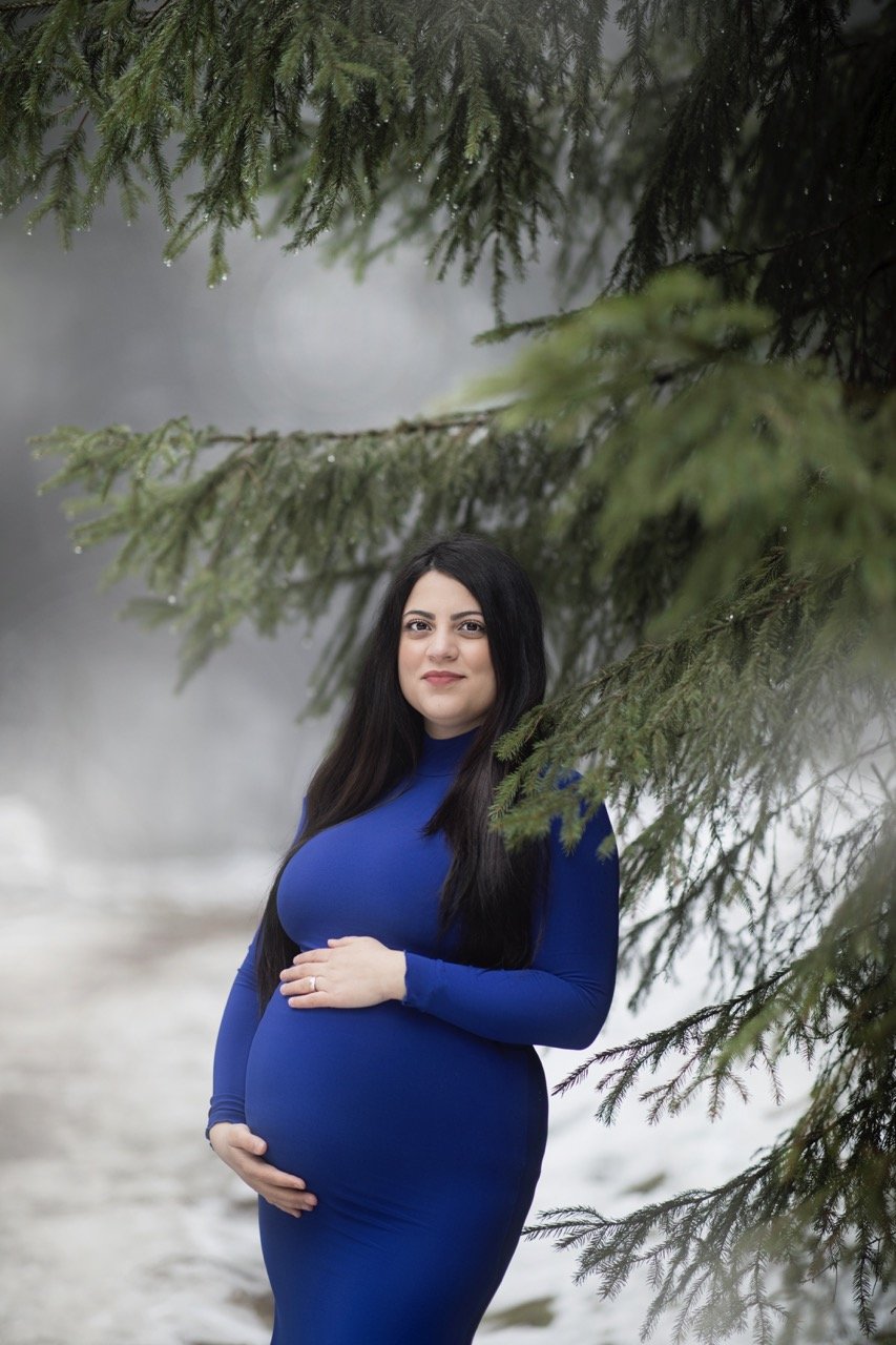 Schwangere Frau in einem blauen Kleid steht unter Tannenzweigen im Freien.