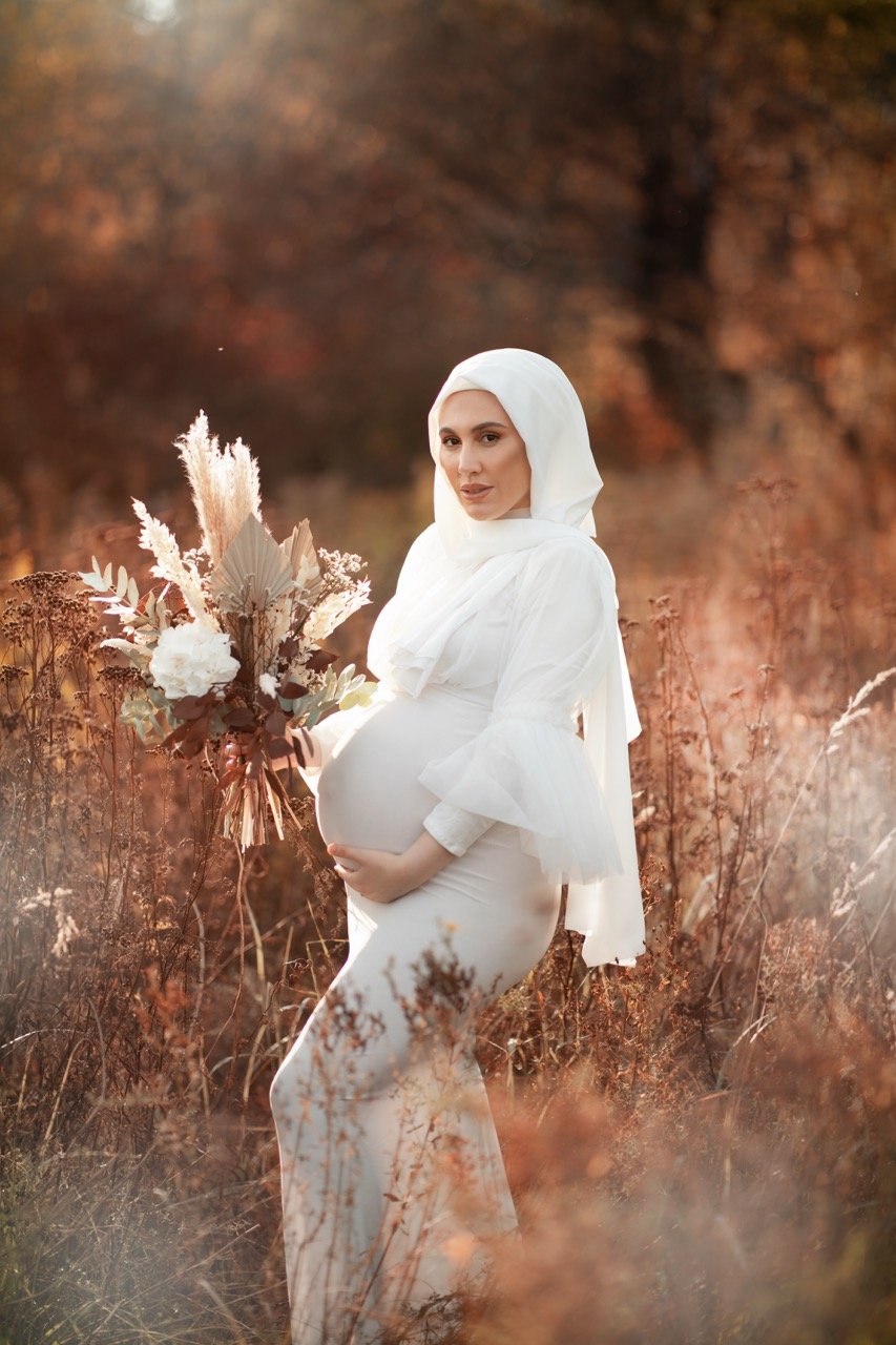 Schwangere Frau im weißen Kleid und Kopftuch hält einen Blumenstrauß in einer herbstlichen Landschaft.