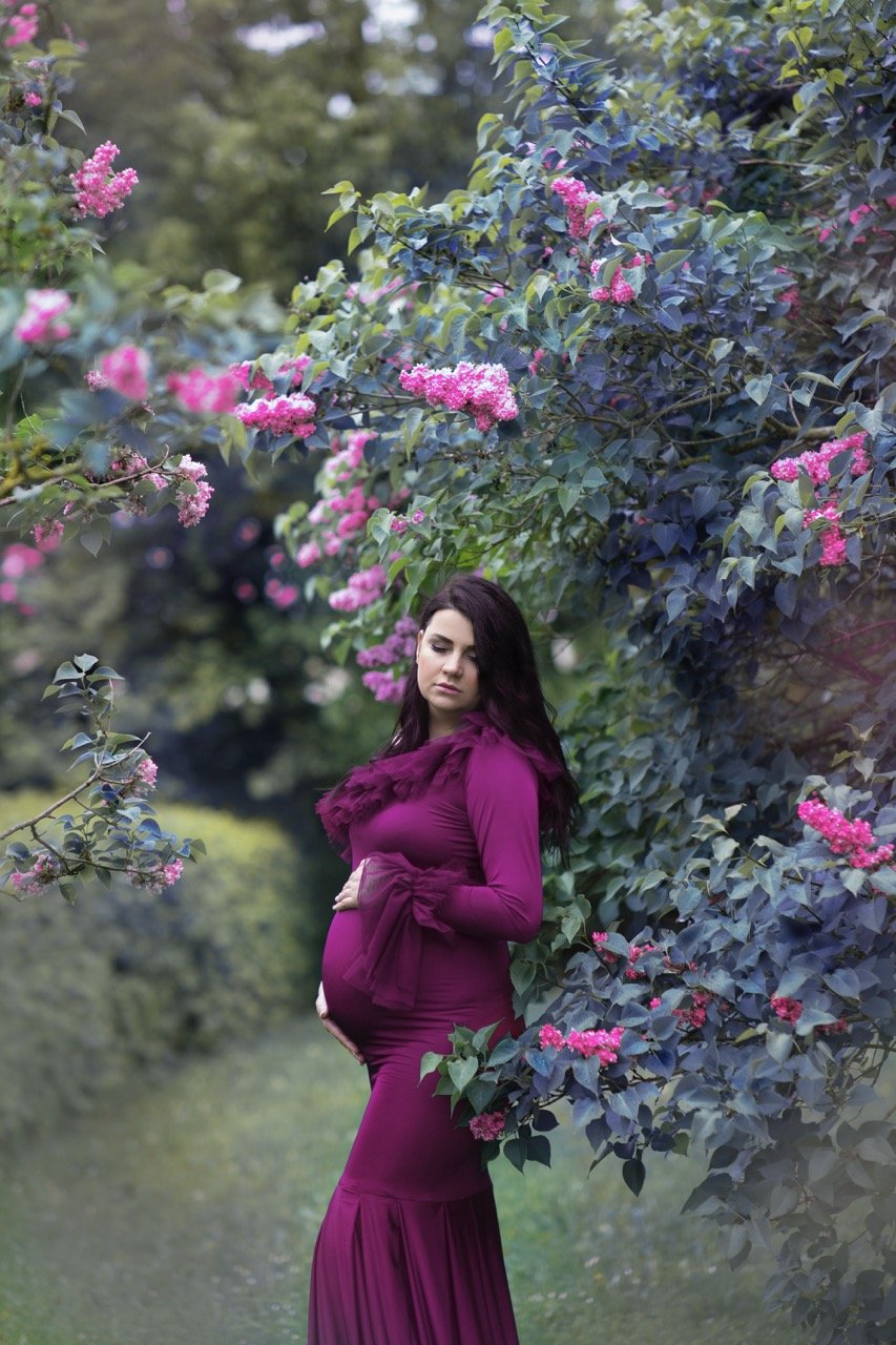 Schwangere Frau in einem lila Kleid steht neben einem blühenden Busch mit rosa Blumen.