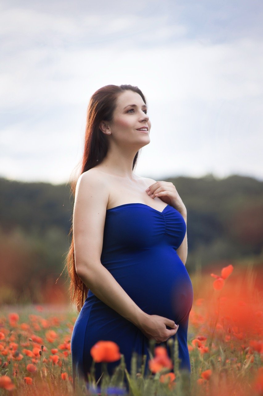 Schwangere Frau in einem blauen Kleid steht auf einem Mohnblumenfeld und schaut in die Ferne.