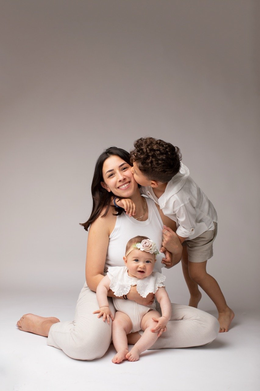 Eine lächelnde Frau sitzt auf dem Boden und hält ein Baby, während ein kleiner Junge ihr einen Kuss auf die Wange gibt. Beide Kinder tragen weiße Kleidung.