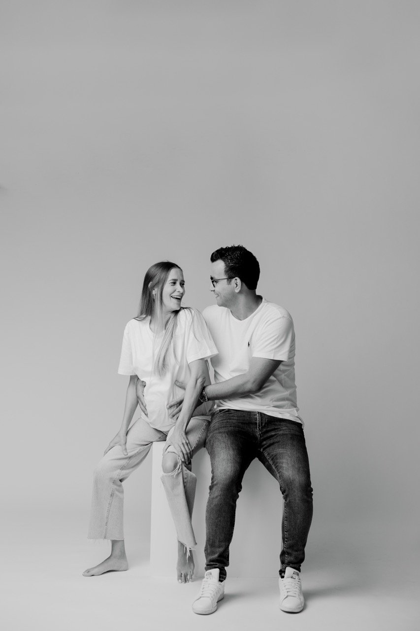 Schwarzweißfoto eines lächelnden Paares, das auf einem Hocker sitzt. Beide tragen helle T-Shirts und Jeans. Der Mann legt einen Arm um die Frau. Sie blicken sich gegenseitig an.