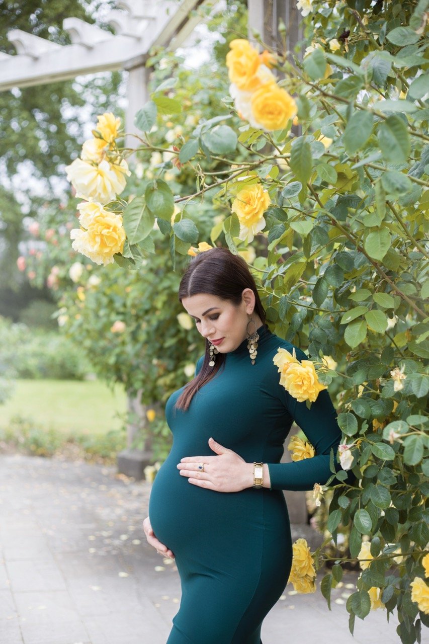 Schwangere Frau steht in einem Garten mit gelben Rosen, trägt ein dunkles, eng anliegendes Kleid.