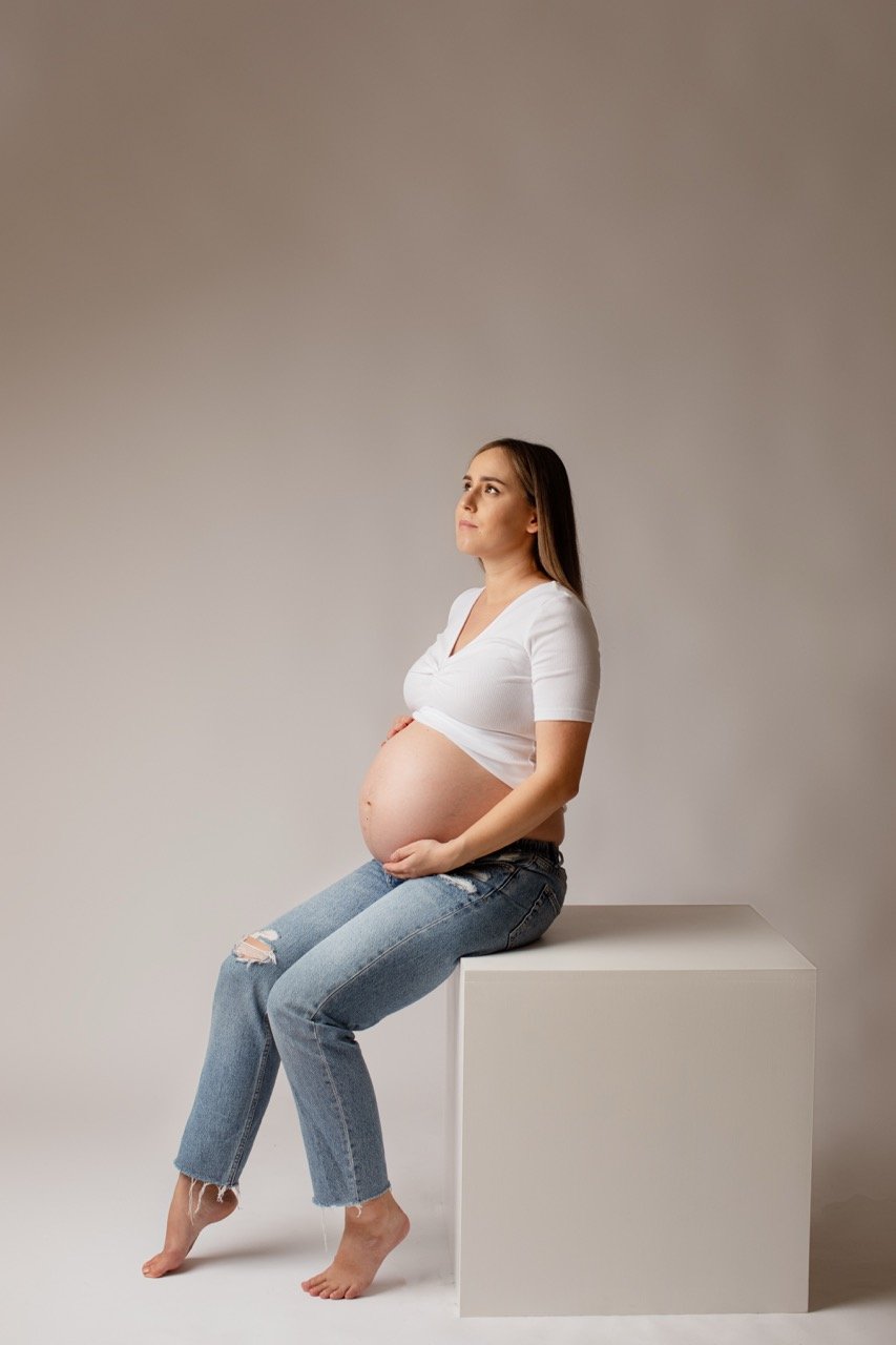 Schwangere Frau sitzt auf einem weißen Würfel, trägt ein weißes Oberteil und Jeans.