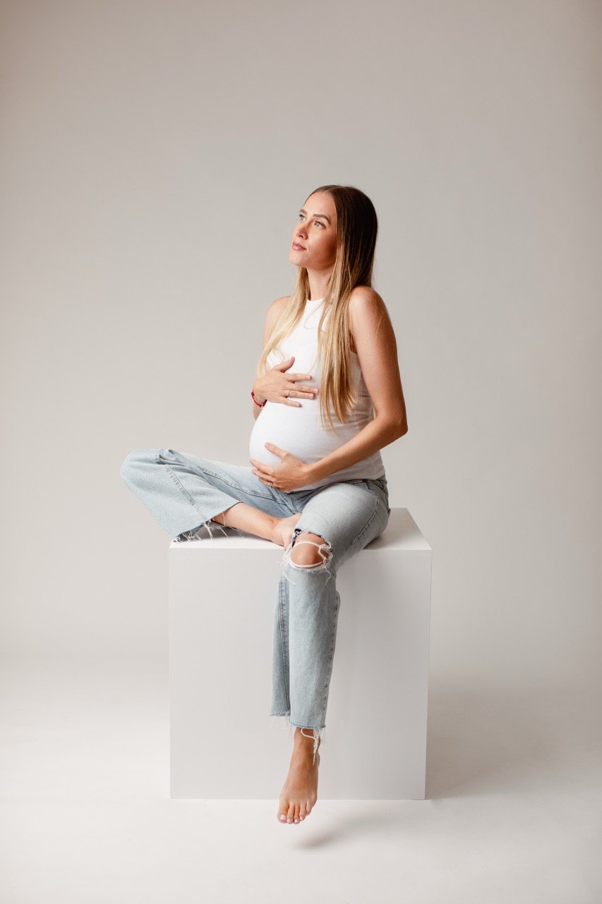 Schwangere Frau in weißem Top und zerrissenen Jeans sitzt auf einem weißen Würfel und hält ihren Bauch.