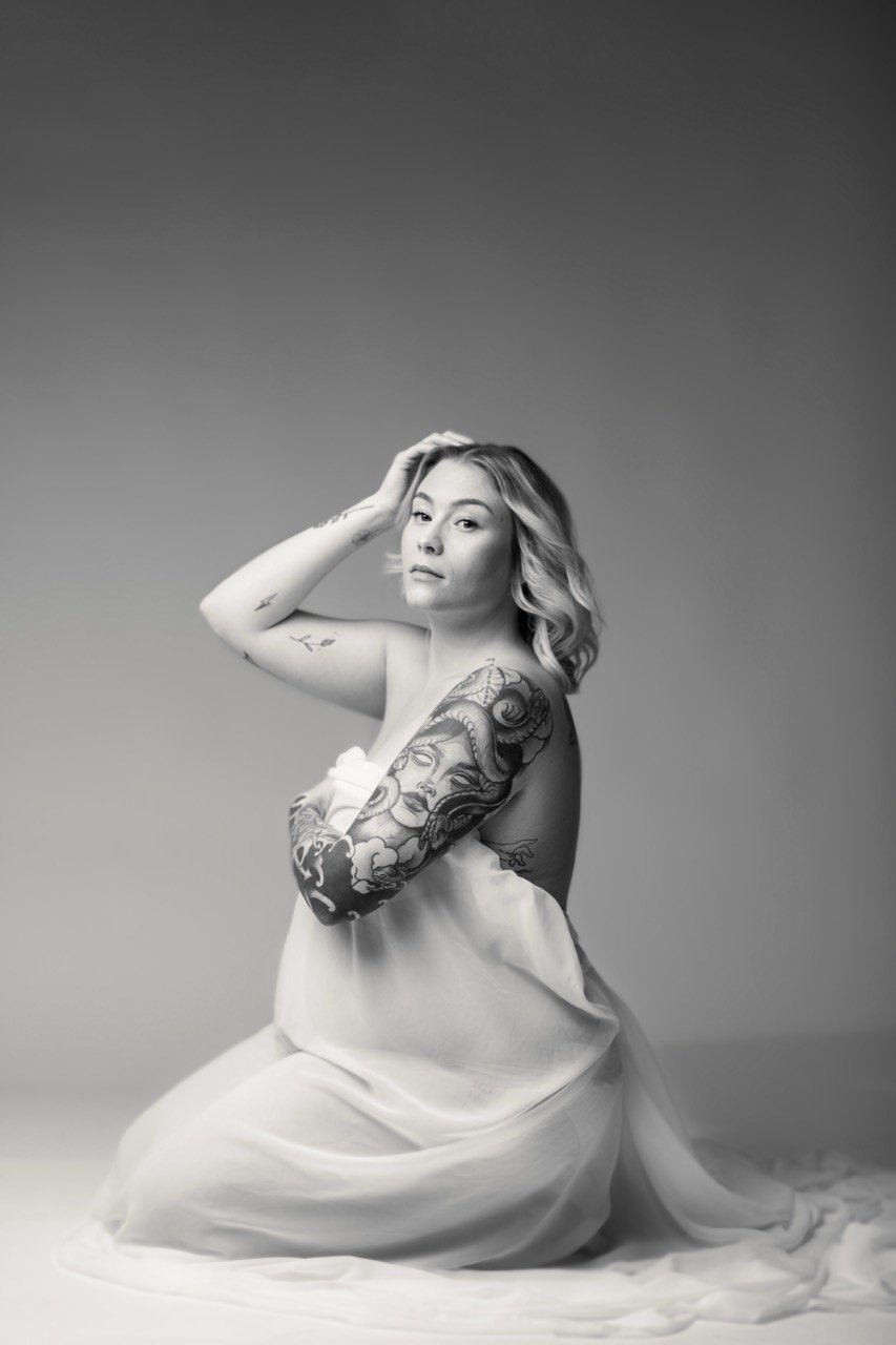 Schwarzweißbild einer Frau mit Tattoos am Arm, die in einem leichten, durchsichtigen Stoff sitzt und in die Kamera schaut.