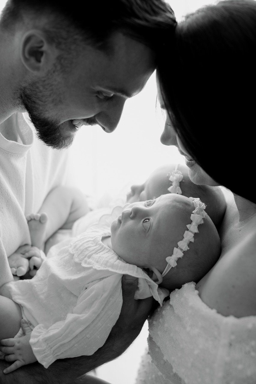 Ein Paar hält ein Baby, das weiße Kleidung und ein Stirnband trägt, in den Armen und lächelt sich an. Das Bild ist in Schwarz-Weiß aufgenommen.