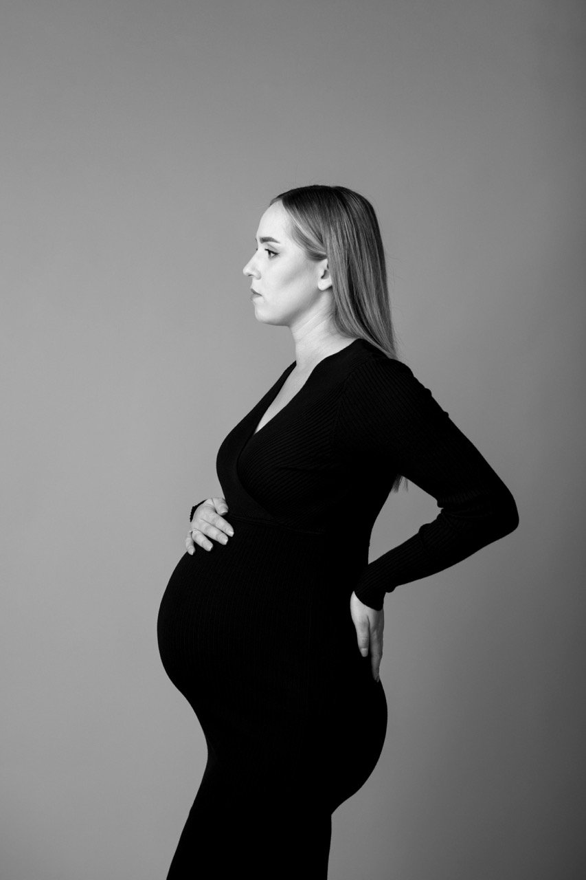 Schwarz-weißes Porträt einer seitlich stehenden, schwangeren Frau in einem schwarzen Kleid, die den Bauch hält.