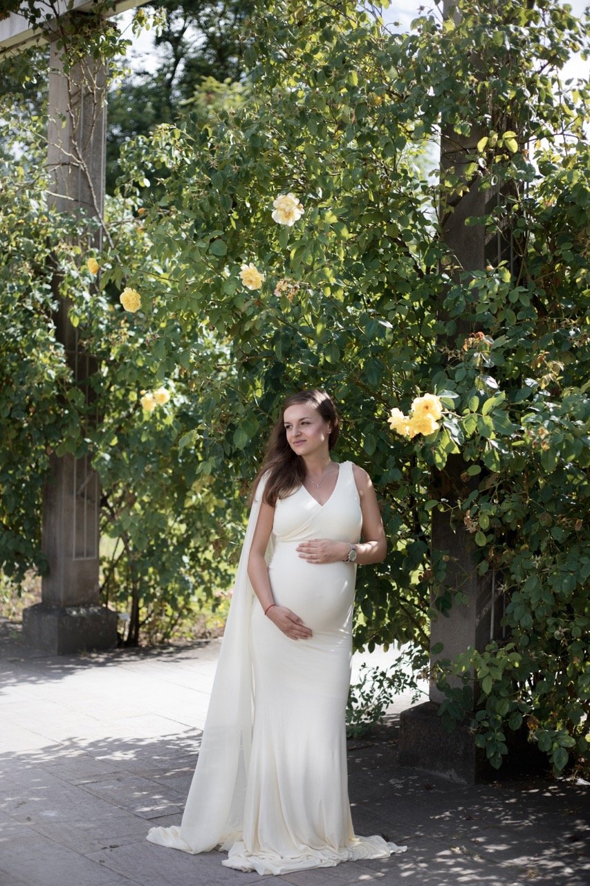 Eine schwangere Frau in einem langen, weißen Kleid steht vor einem blühenden Rosenstrauch.