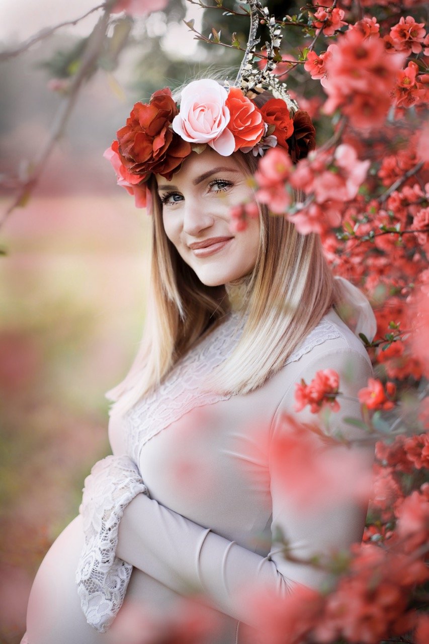 Schwangere Frau mit Blumenkranz posiert lächelnd vor blühenden roten Blumen im Freien.