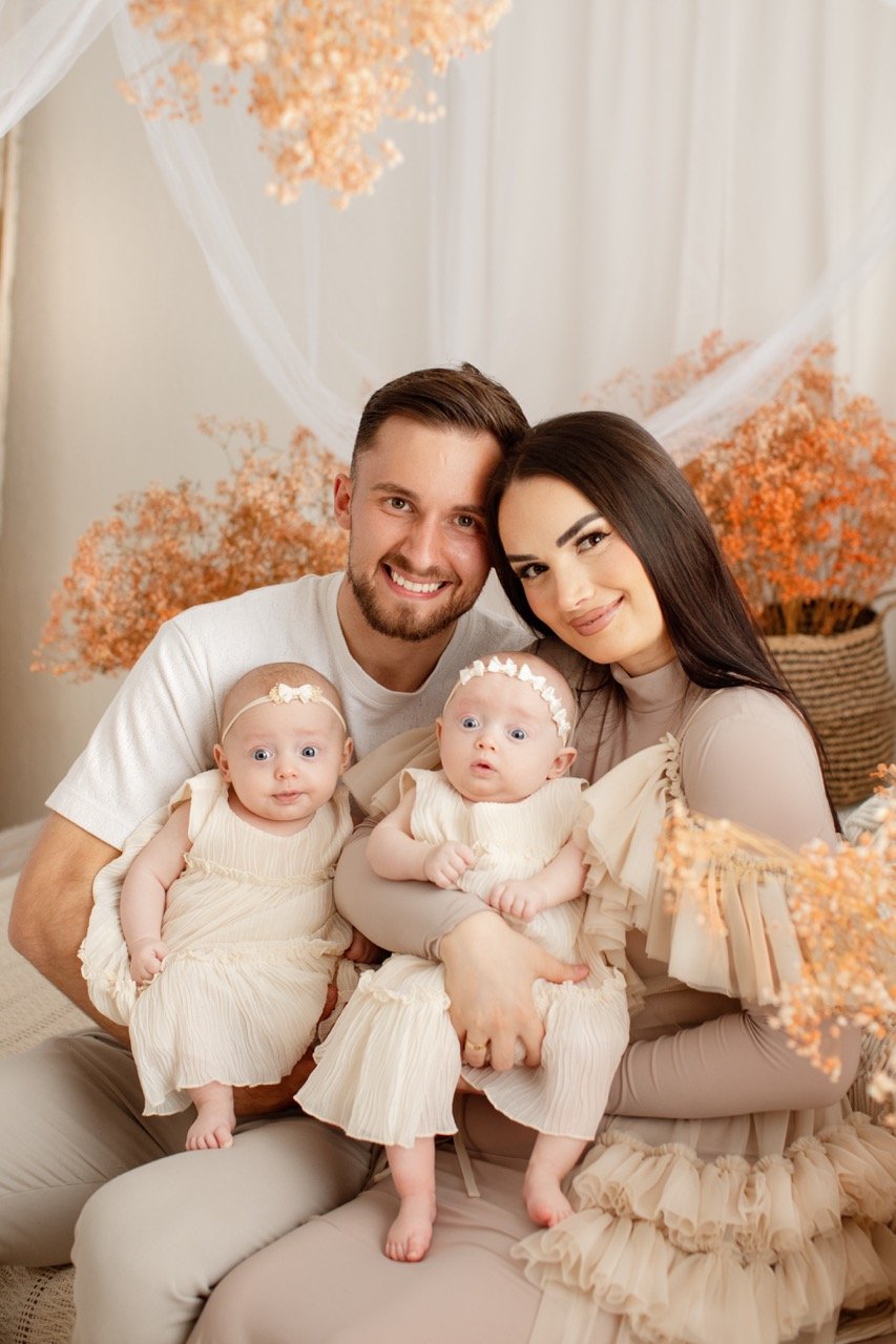 Ein lächelndes Paar, das zwei Babys in cremefarbener Kleidung hält, sitzt vor einem Hintergrund mit orangefarbenen Trockenblumen.