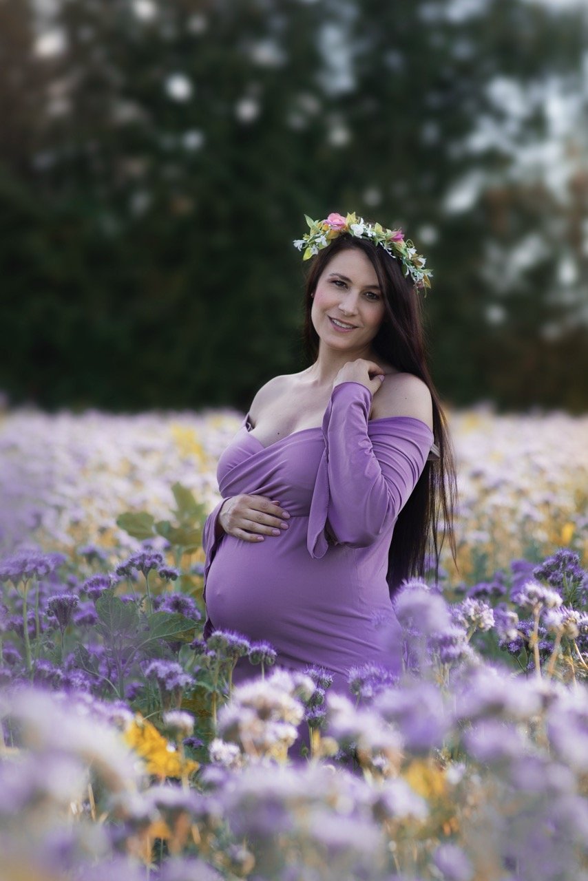 Schwangere Frau im lila Kleid steht in einem Blumenfeld, trägt einen Blumenkranz im Haar.