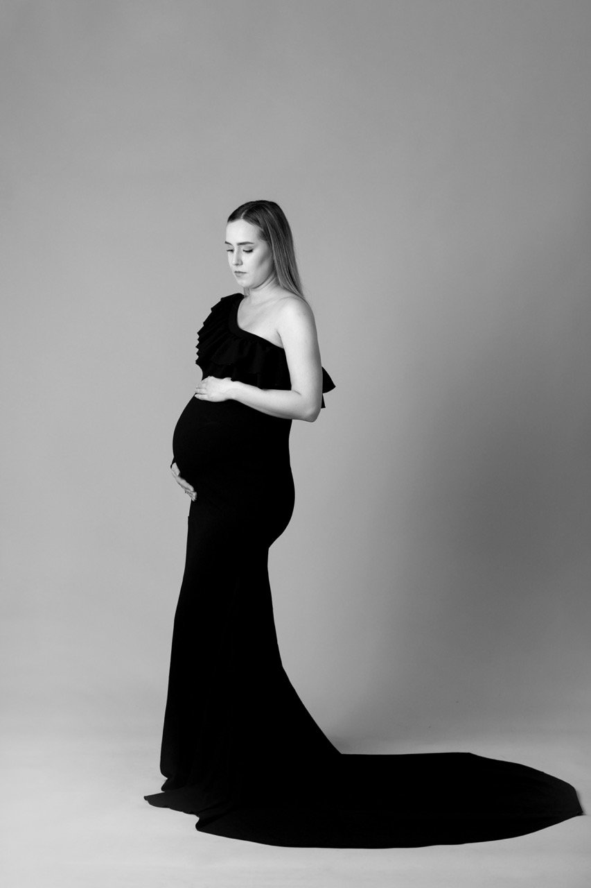Schwarz-Weiß-Foto einer schwangeren Frau in einem eleganten, langen Kleid, die ihren Babybauch hält.